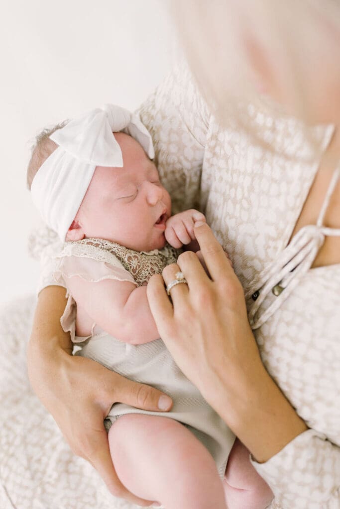 Close-up of a newborn baby girl in a lace outfit and white bow, cradled in her mother’s arms during a soft and timeless newborn session.