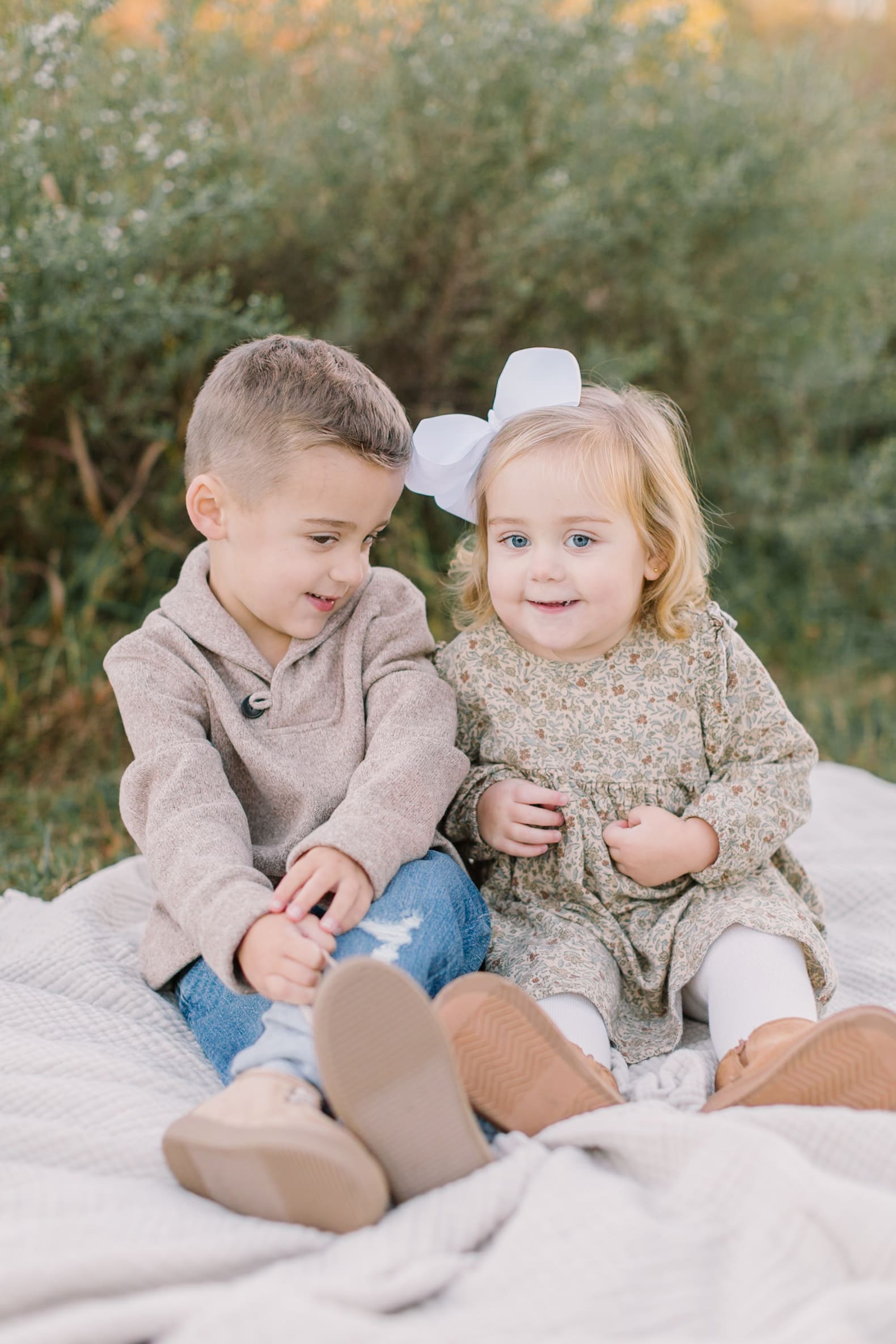Toddler brother and sister play together on a picnic blanket in a park with tall grasses