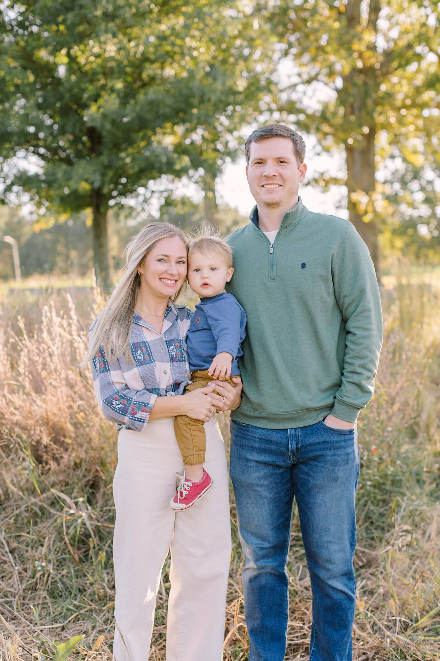 Smiling mom and dad stand in a park at sunset with their toddler on mom's hip