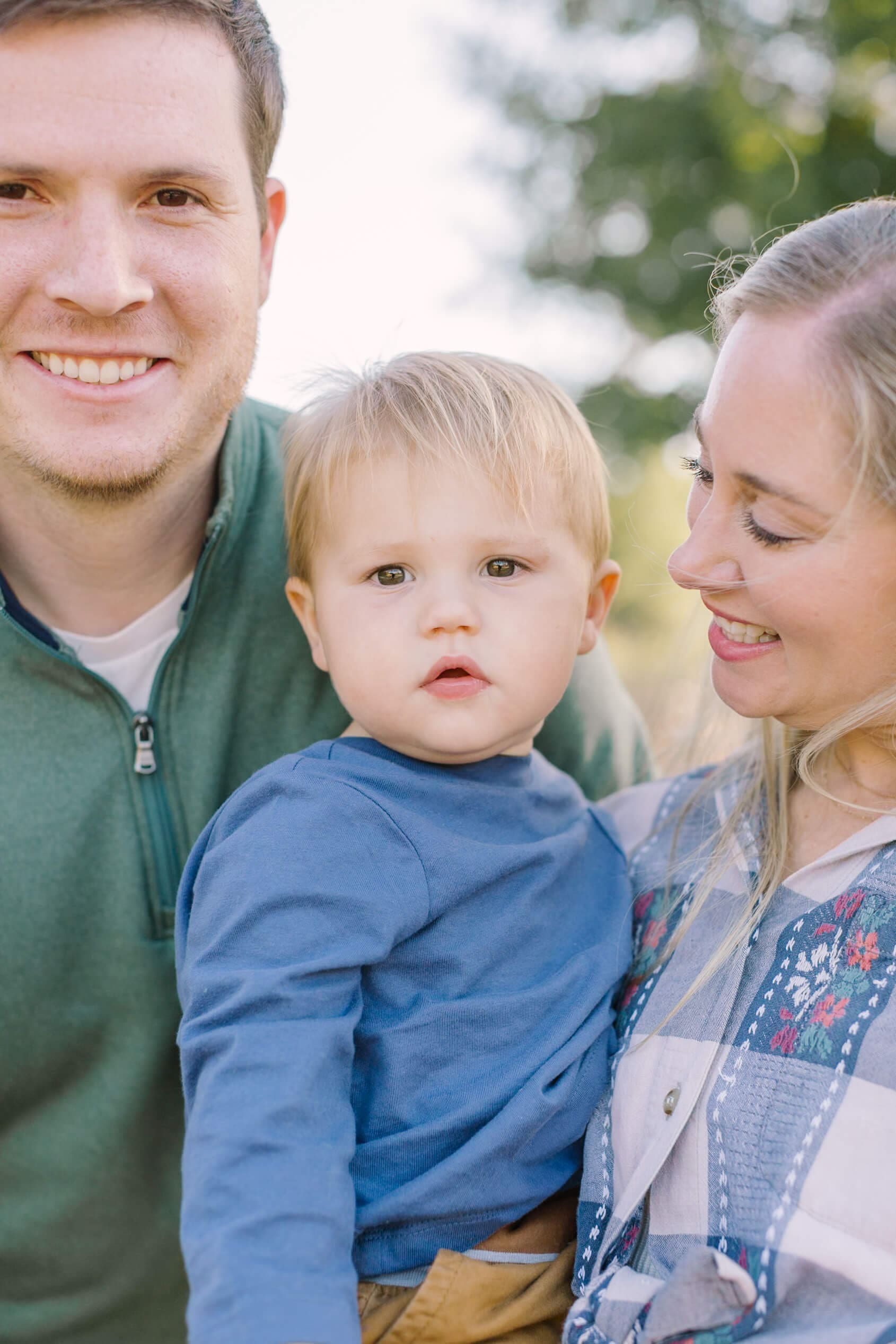 A toddler boy in a blue shirt sits in mom's arms as they stand in a park with happy dad