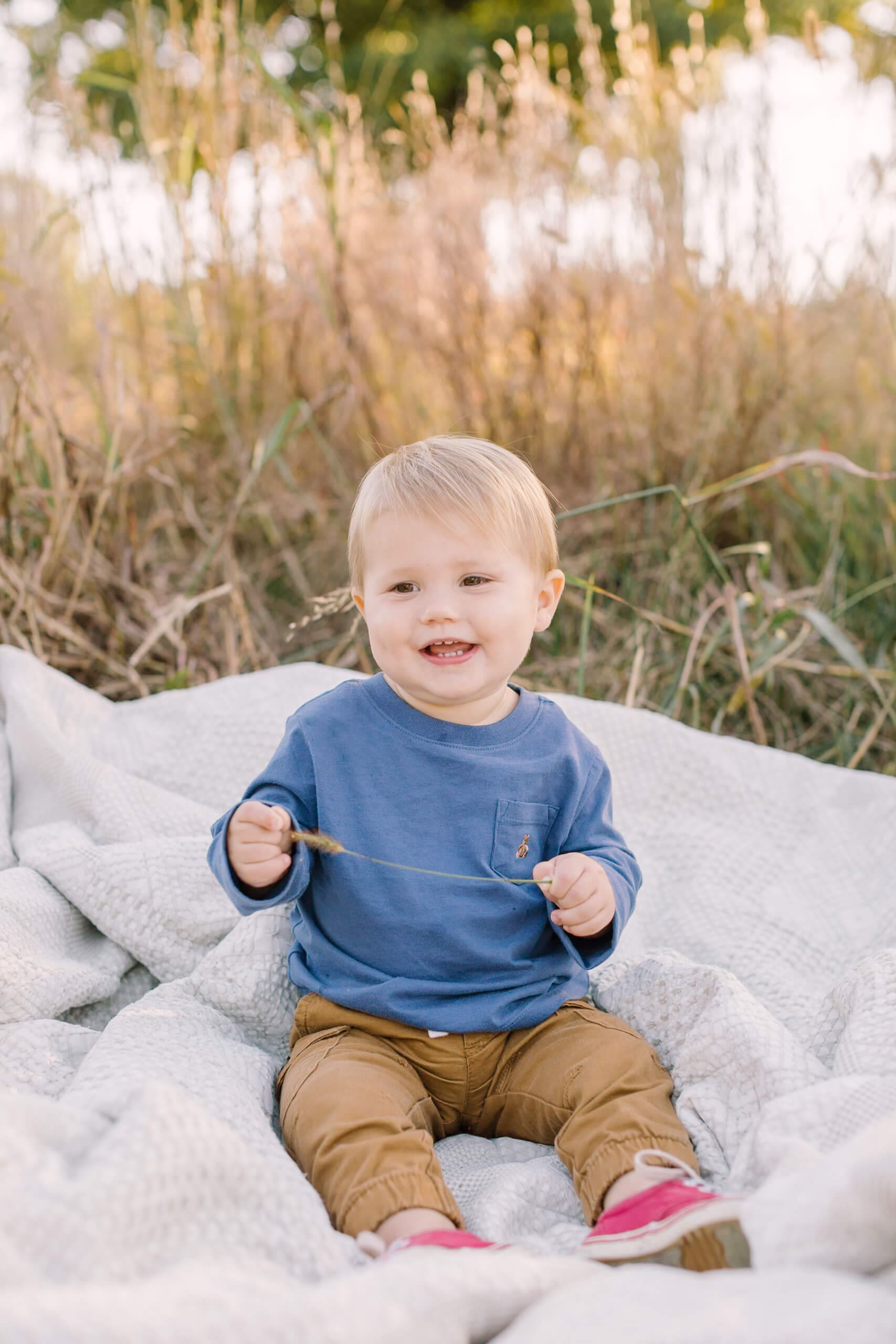 A toddler boy in a blue shirt sits on a blanket in a park lawn with tall grass after visiting christmas tree farms in raleigh