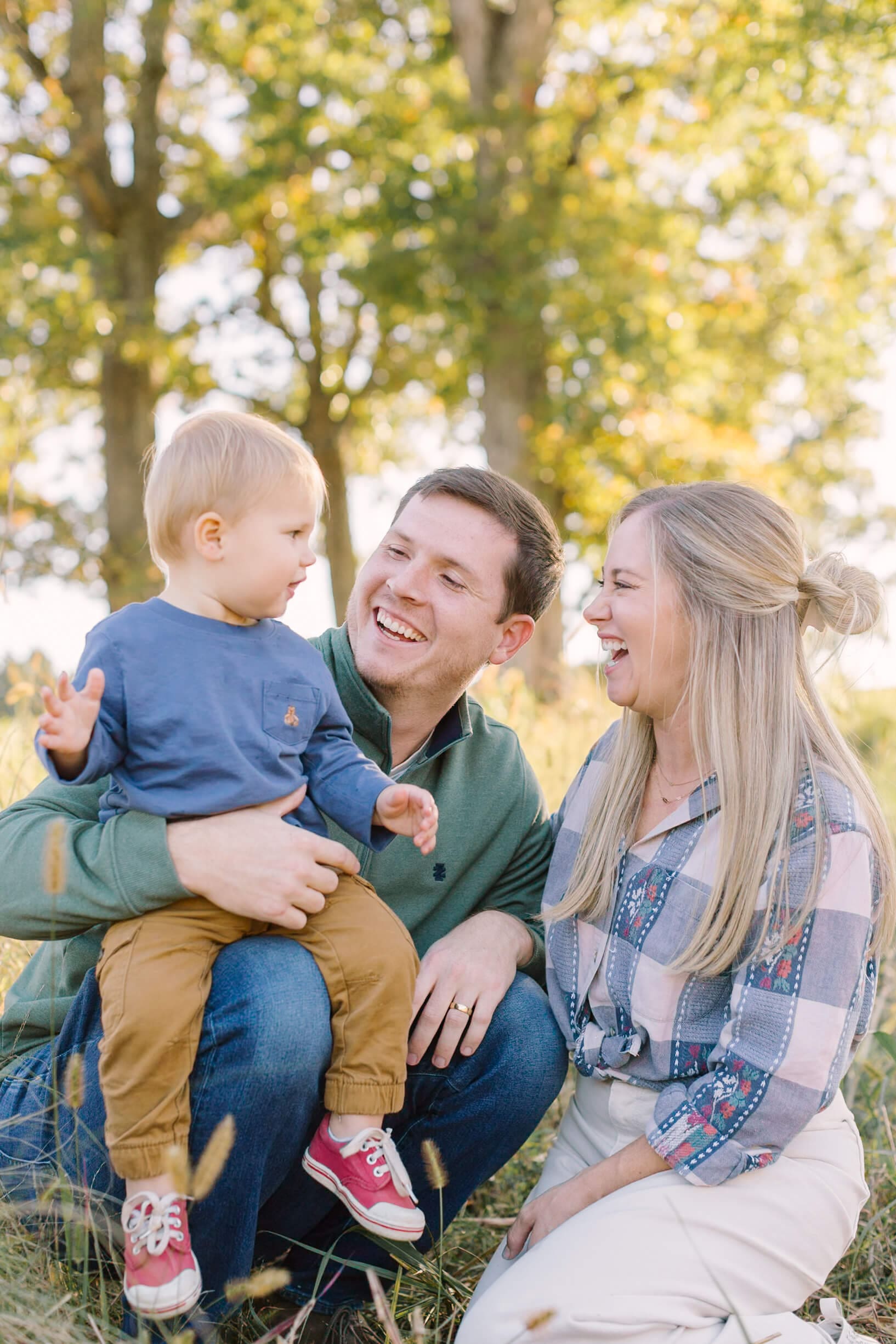 A happy mom and dad laugh with their toddler son on dad's knee while playing in a lawn with tall grass at sunset after visiting christmas tree farms in raleigh