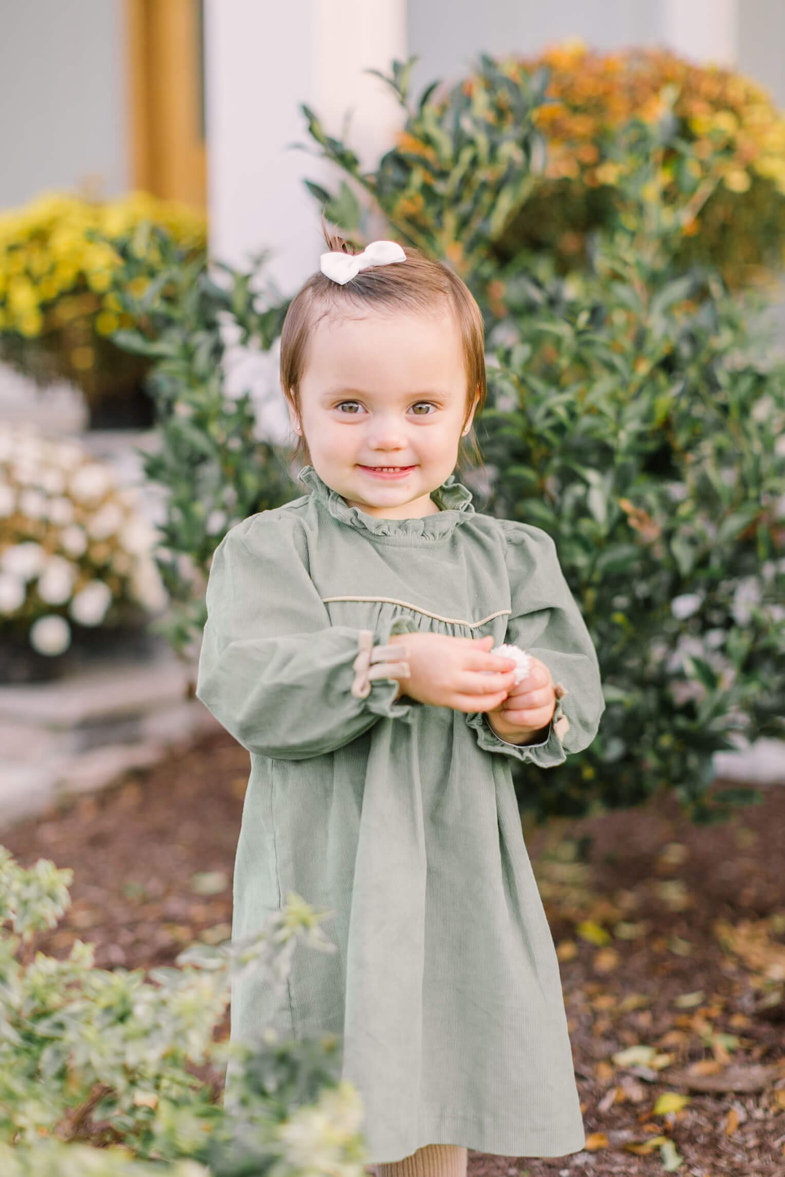 A toddler girl in a green dress explores flowers in the garden with a smile before visiting fall festivals in raleigh