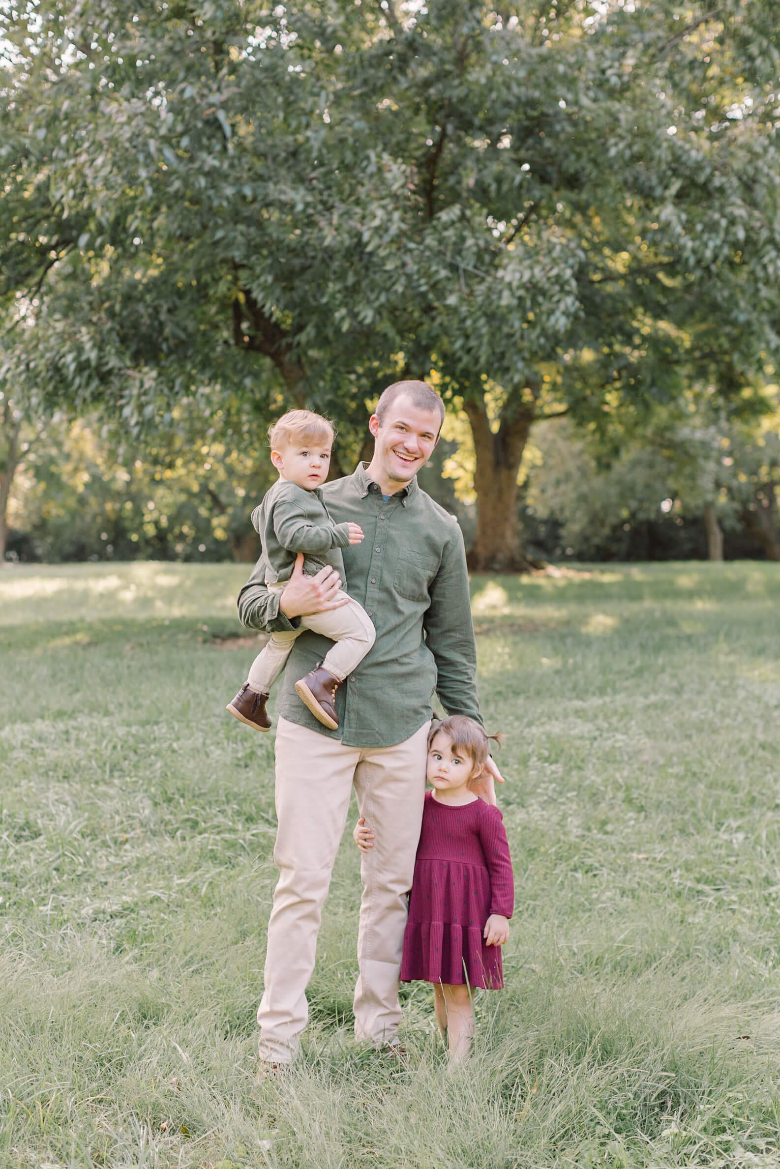 A happy dad stands with his young daughter and son on his hip in a lawn