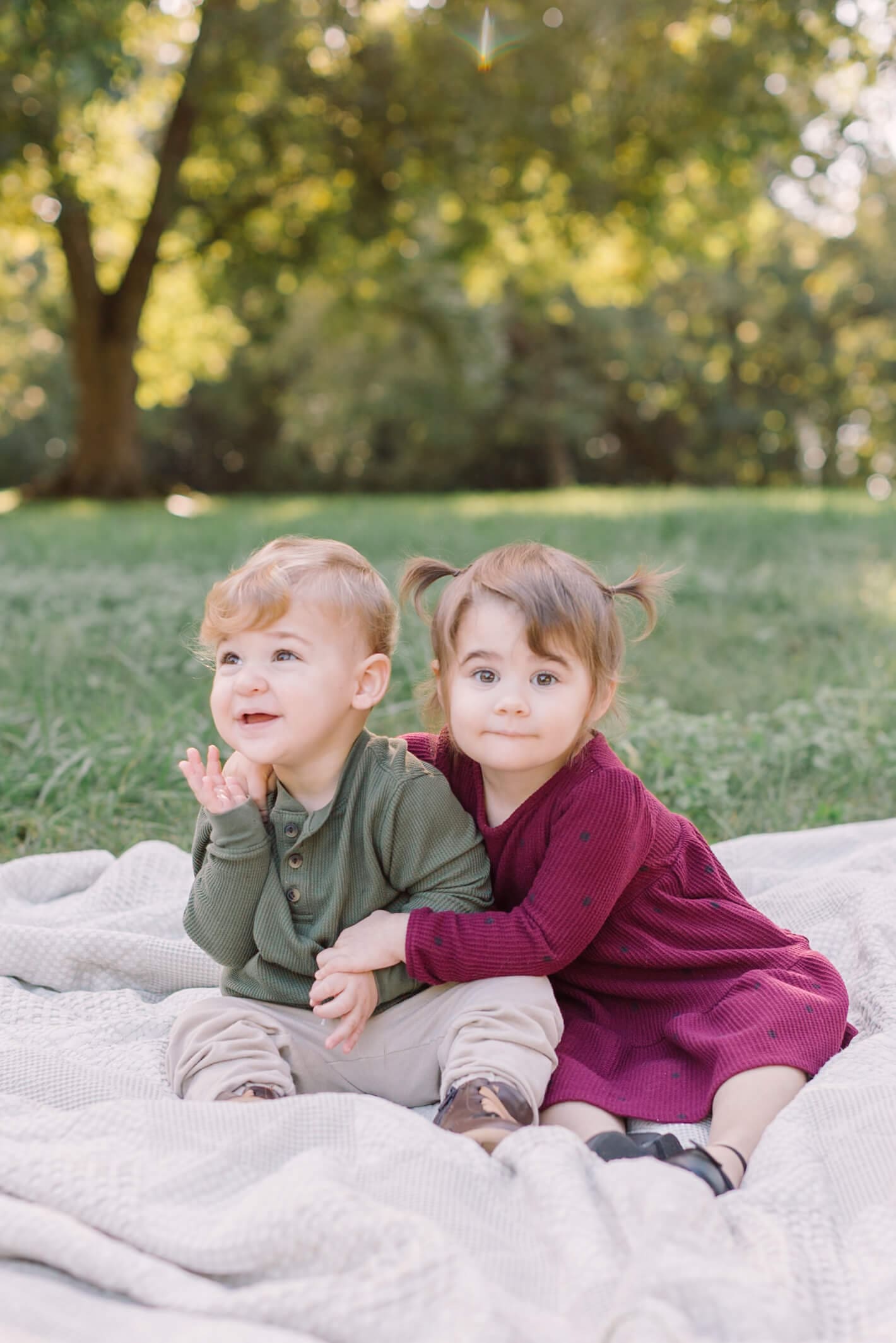 A toddler girl in a purple dress hugs her baby brother as they sit on a picnic blanket before visiting a pumpkin patch in raleigh