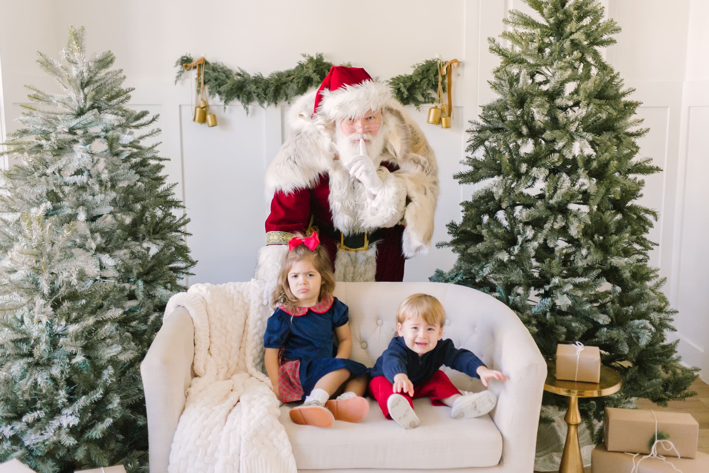 Two young toddler sit on a couch in front of saint nick in a studio
