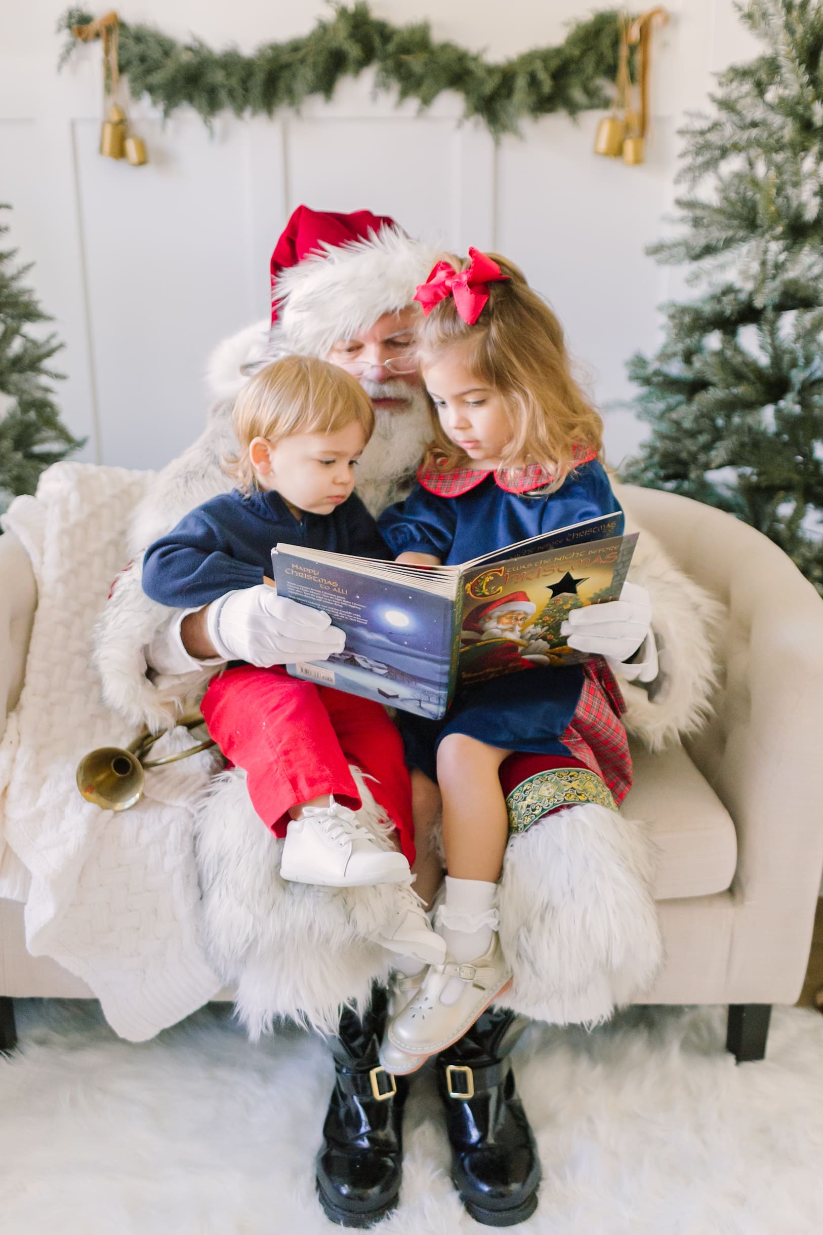 Santa reads a christmas book to two toddlers in his lap on a couch