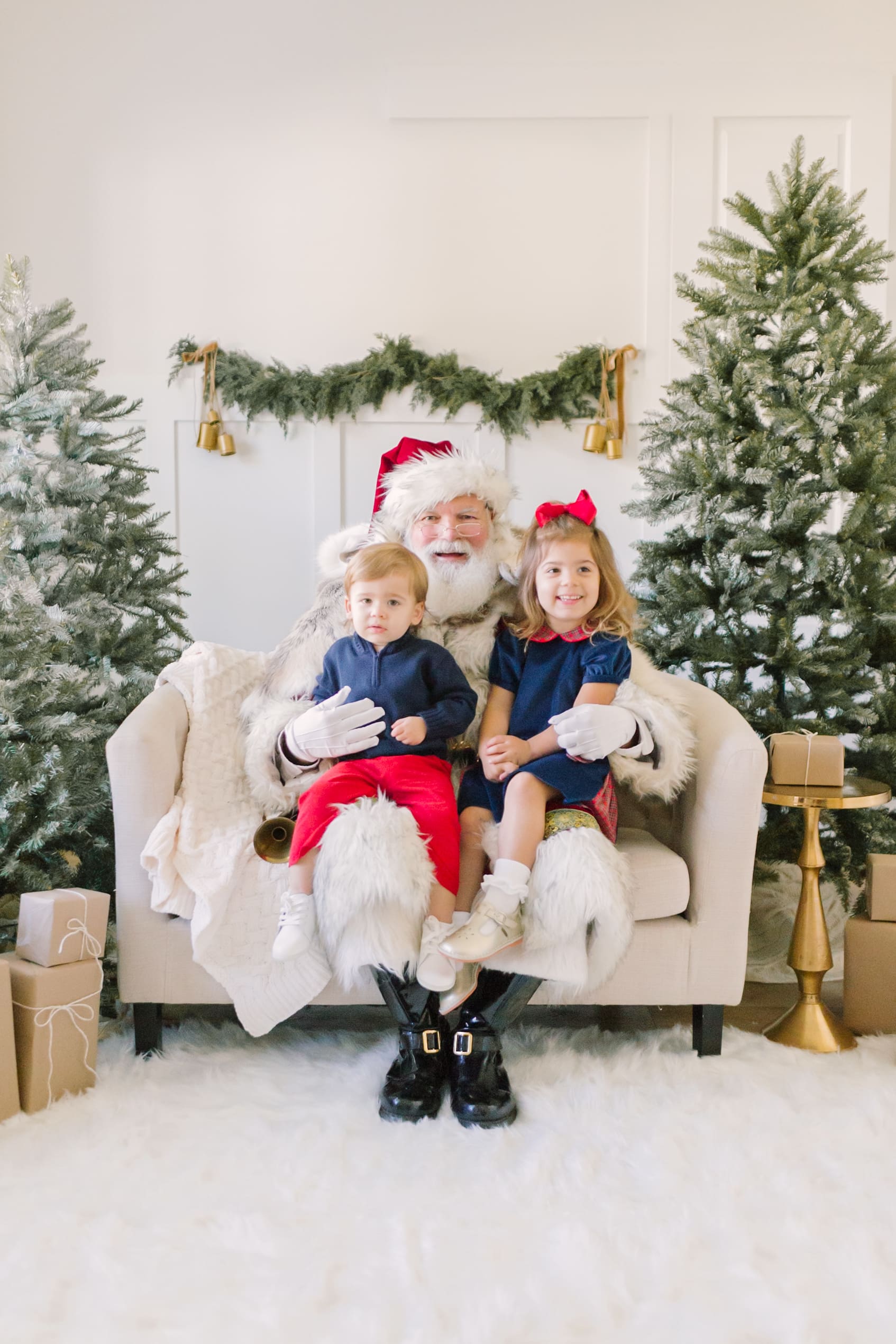 A toddler girl and her younger brother smile while sitting on the lap of a santa in raleigh wearing matching blue outfits