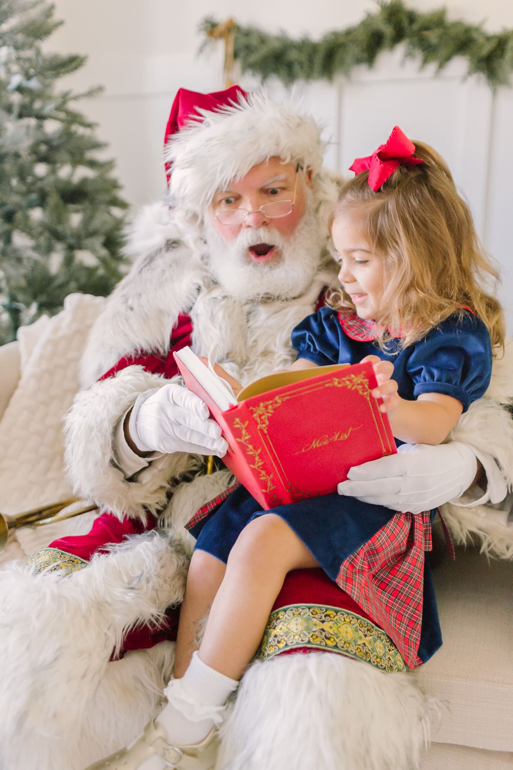 A toddler girl in a blue dress reads a book with a santa in raleigh while sitting on his lap in a studio
