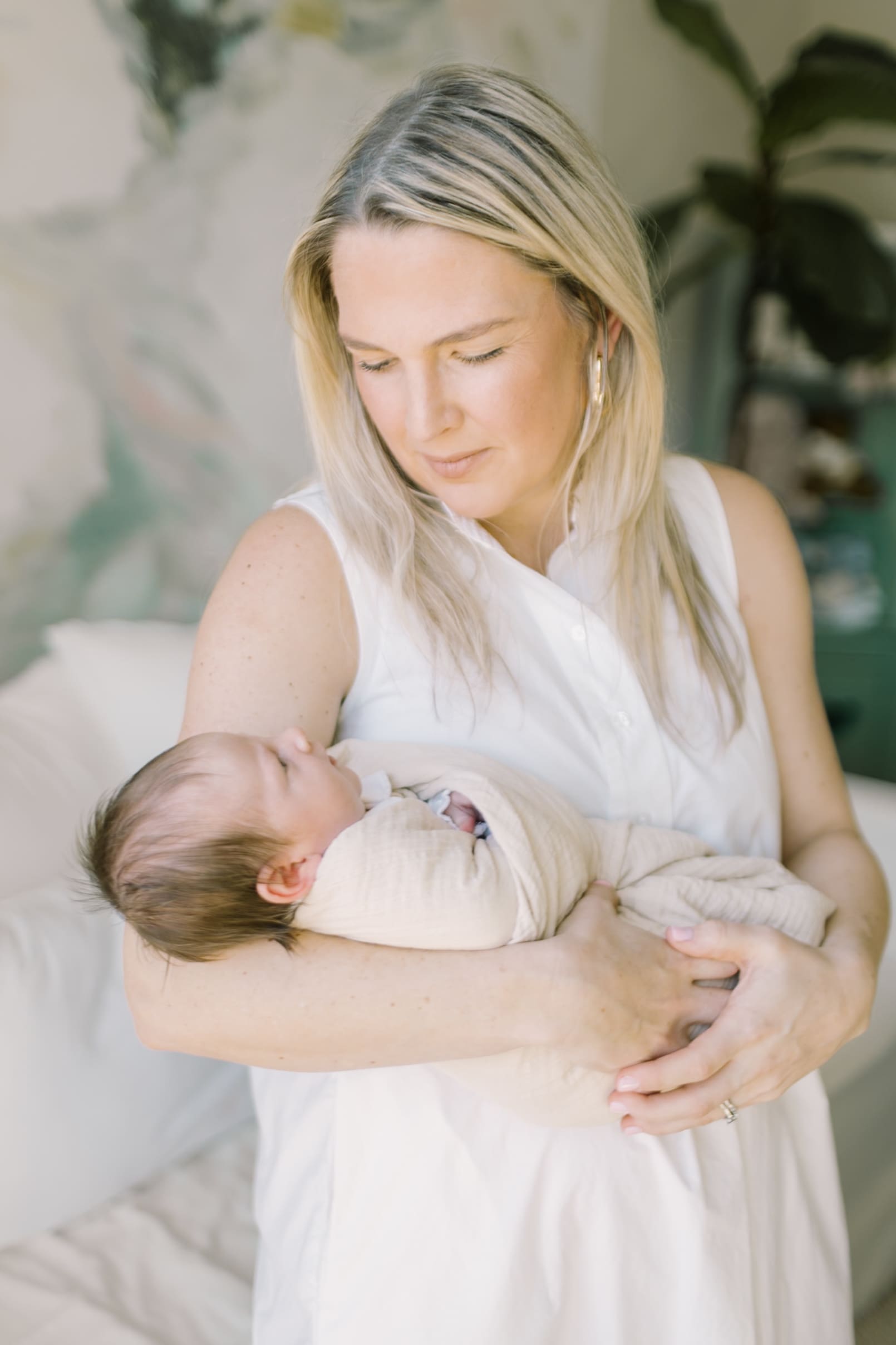 A new mom in a white dress looks down to her newborn laying in her arms in a tan swaddle