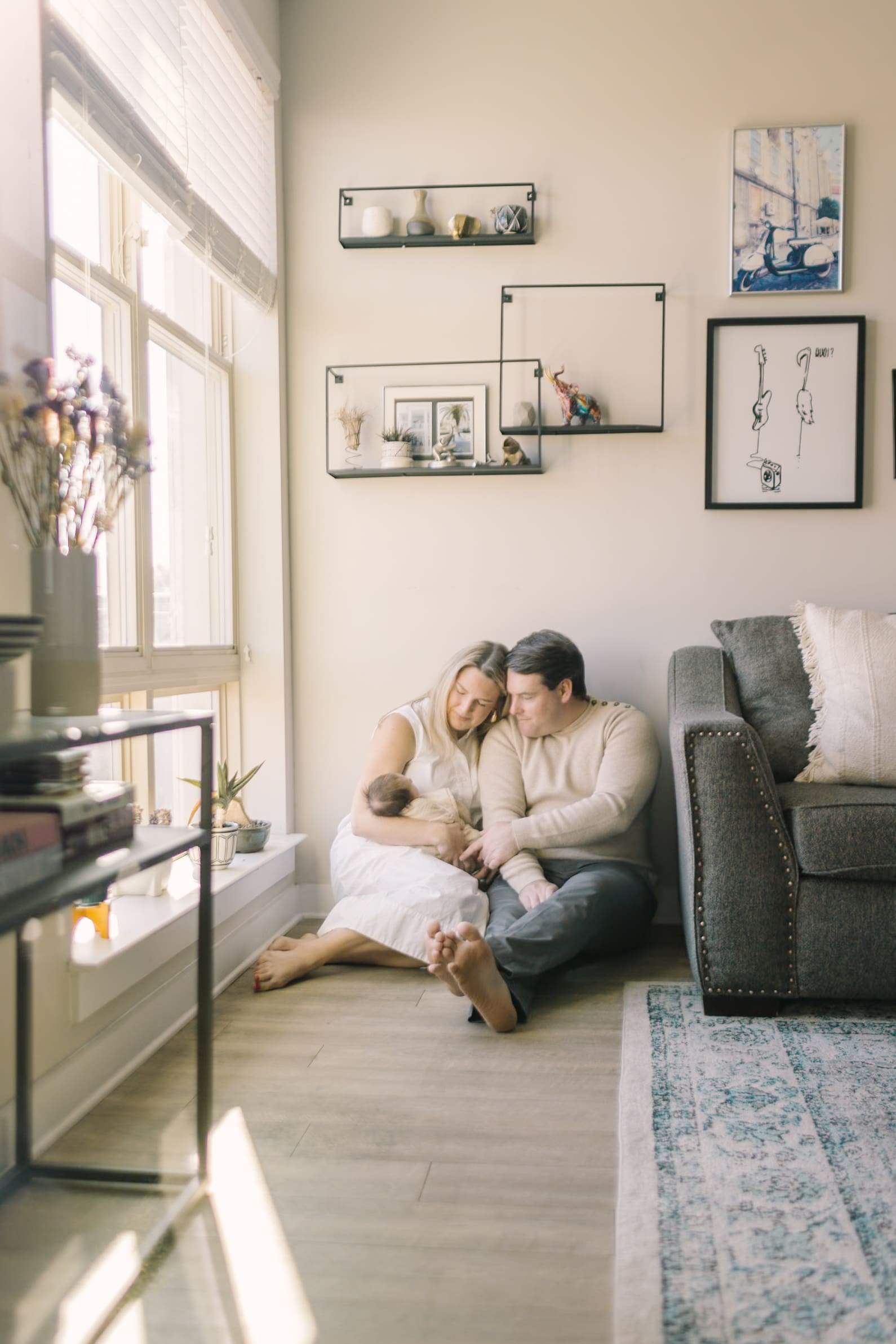 New parents sit in their living room floor under a window cradling their sleeping newborn daughter
