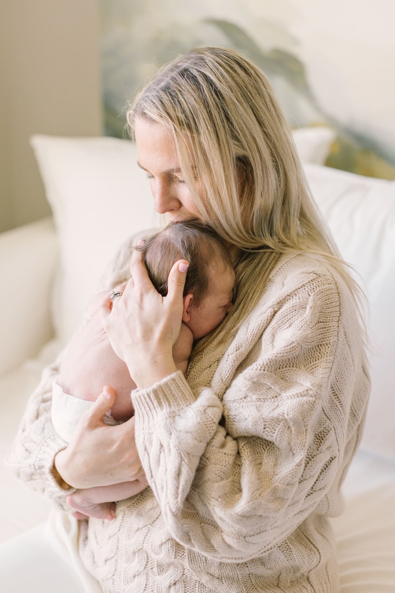 A new mom in a tan knit sweater cradles her newborn against her chest on a couch after meeting lactation consultants in Raleigh, NC