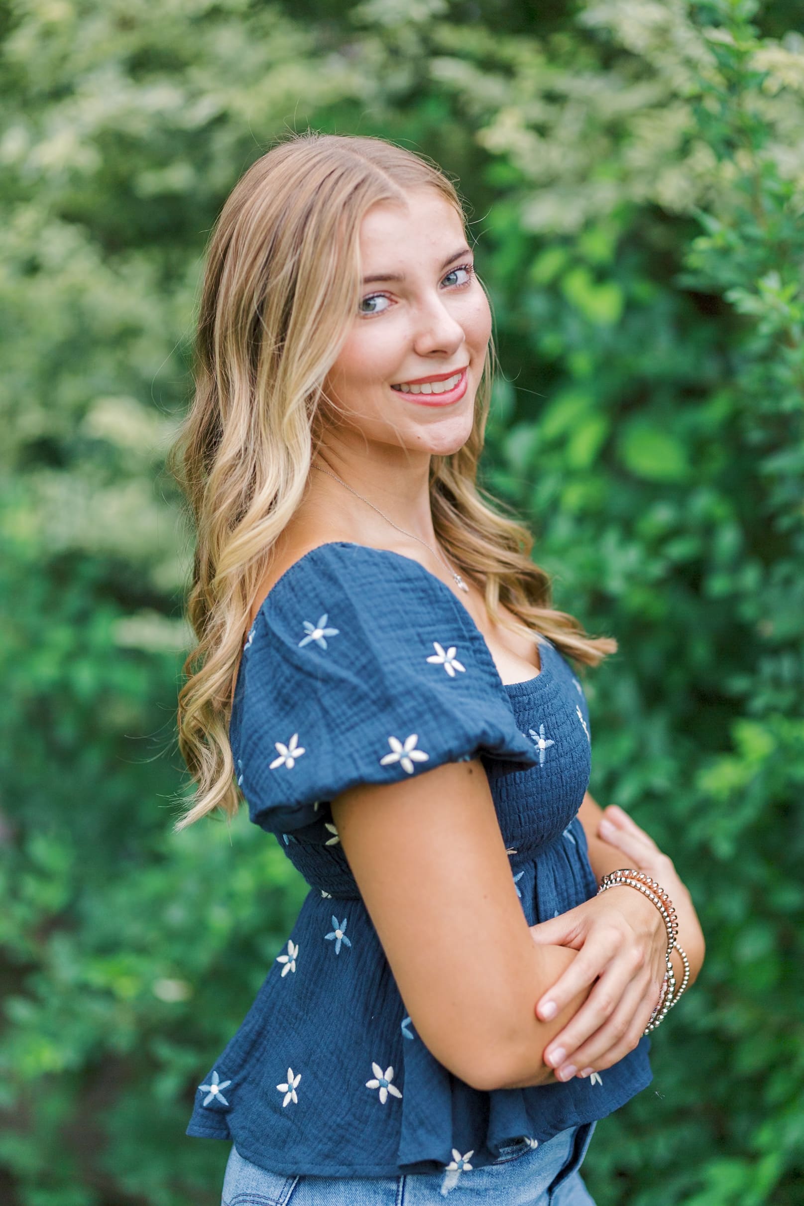A high school senior stands in a blue blouse in a park with arms crossed smiling over her shoulder