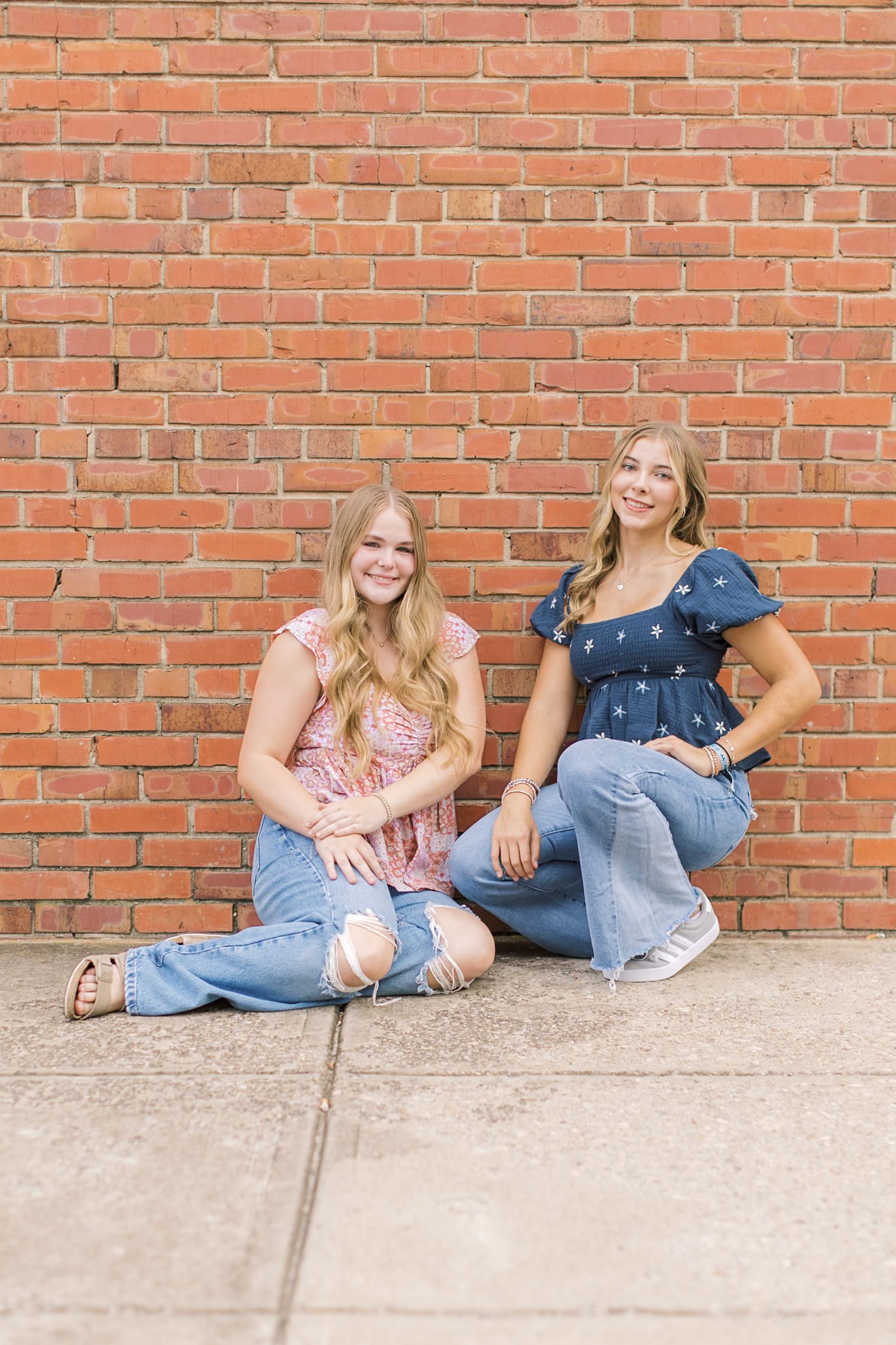 High school best friends in jeans and pink and blue blouses sit against a brick wall smiling after some Raleigh SAT prep
