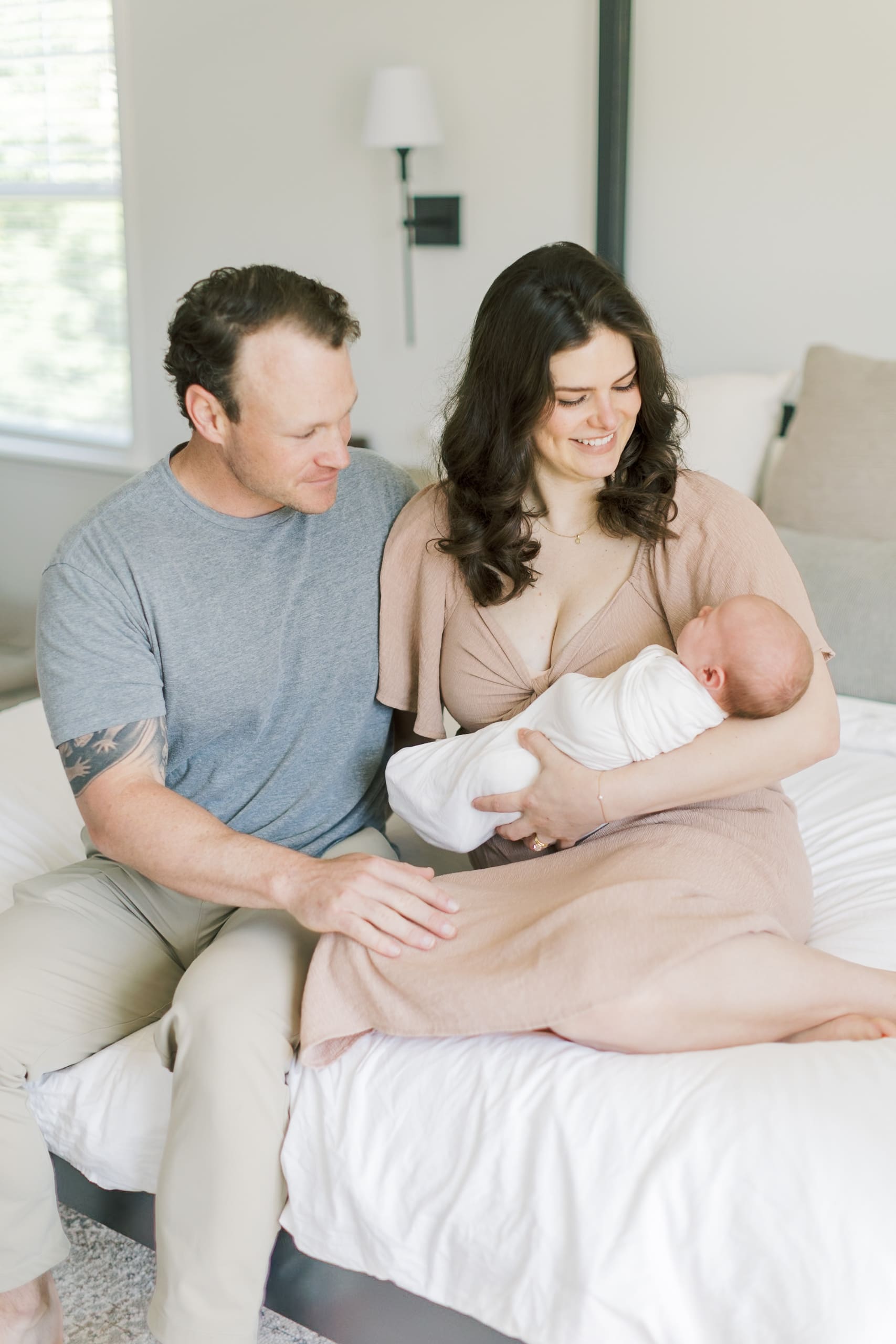 Happy mom and dad sit on a bed together smiling down to their sleeping newborn in mom's arms