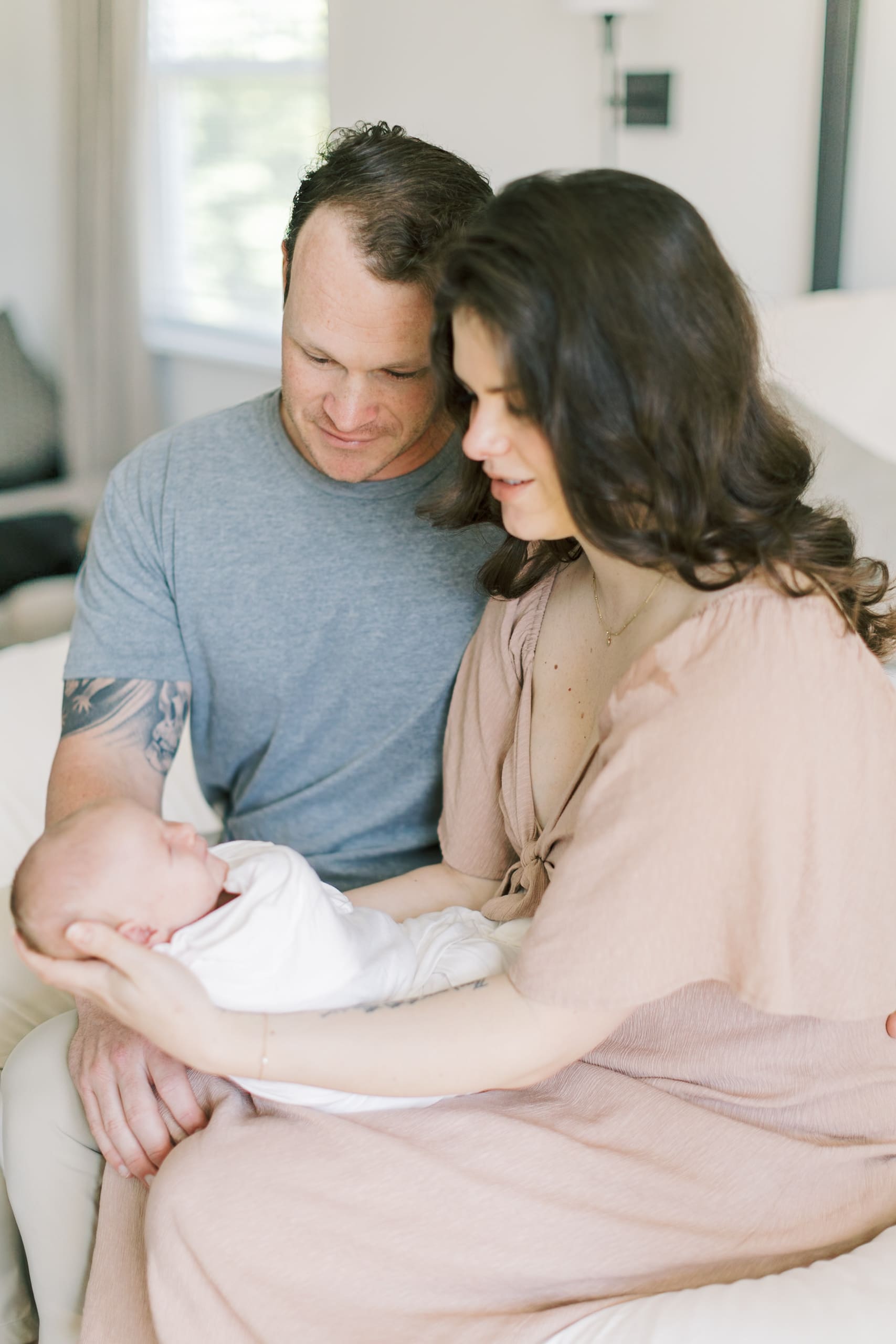 Smiling new parents admire their sleeping newborn in mom's lap on a couch