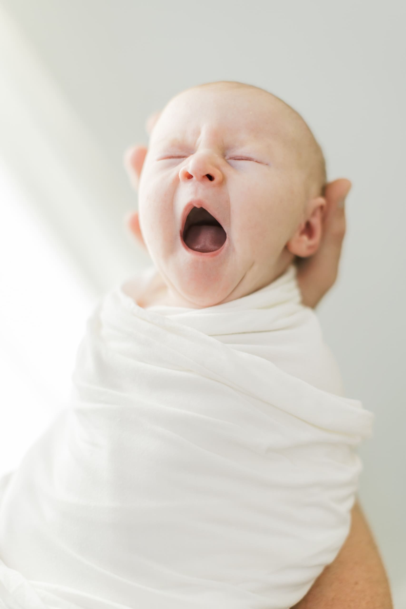 A newborn baby yawns while in dad's hands in a white swaddle after visiting Raleigh baby stores