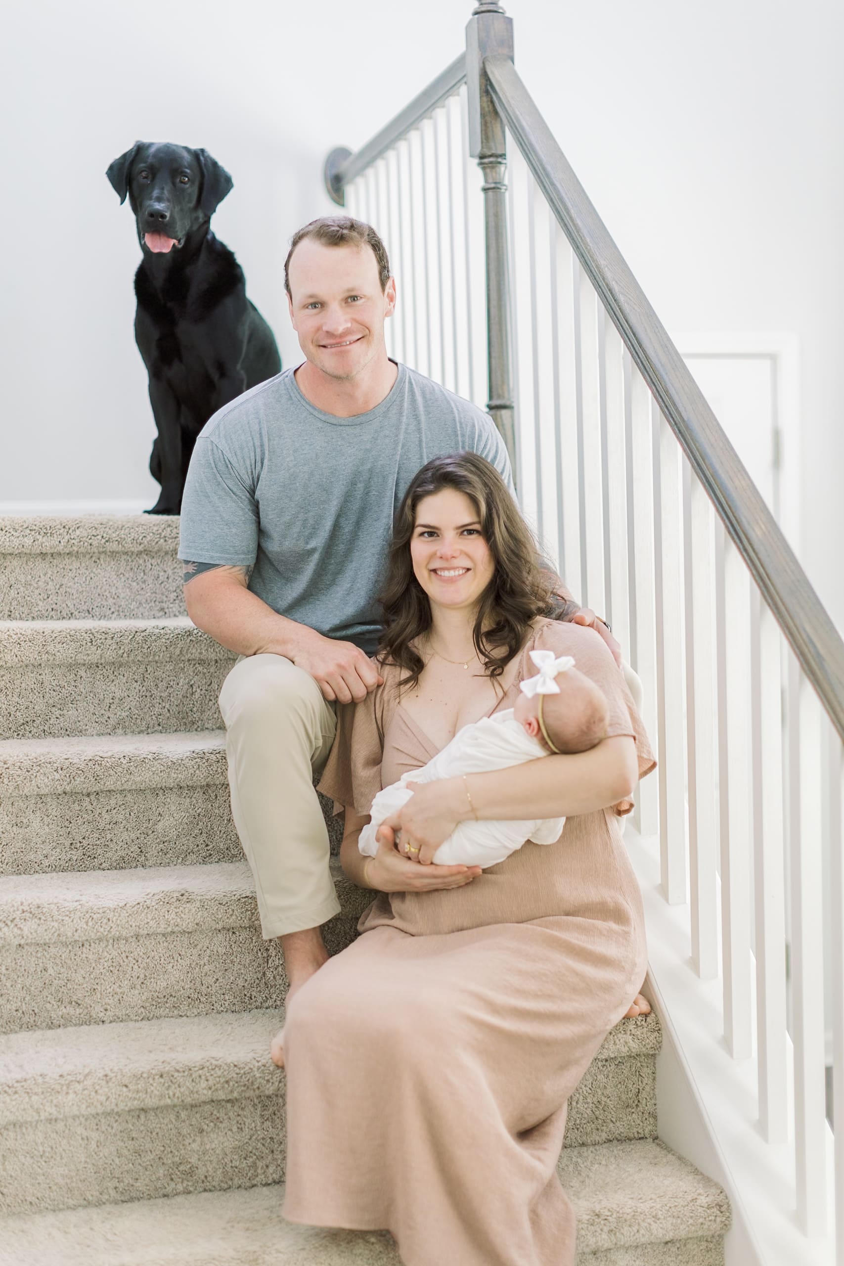 A happy mom and dad sit on their stairs with their newborn in mom's arms and black dog above them after visiting Raleigh baby stores