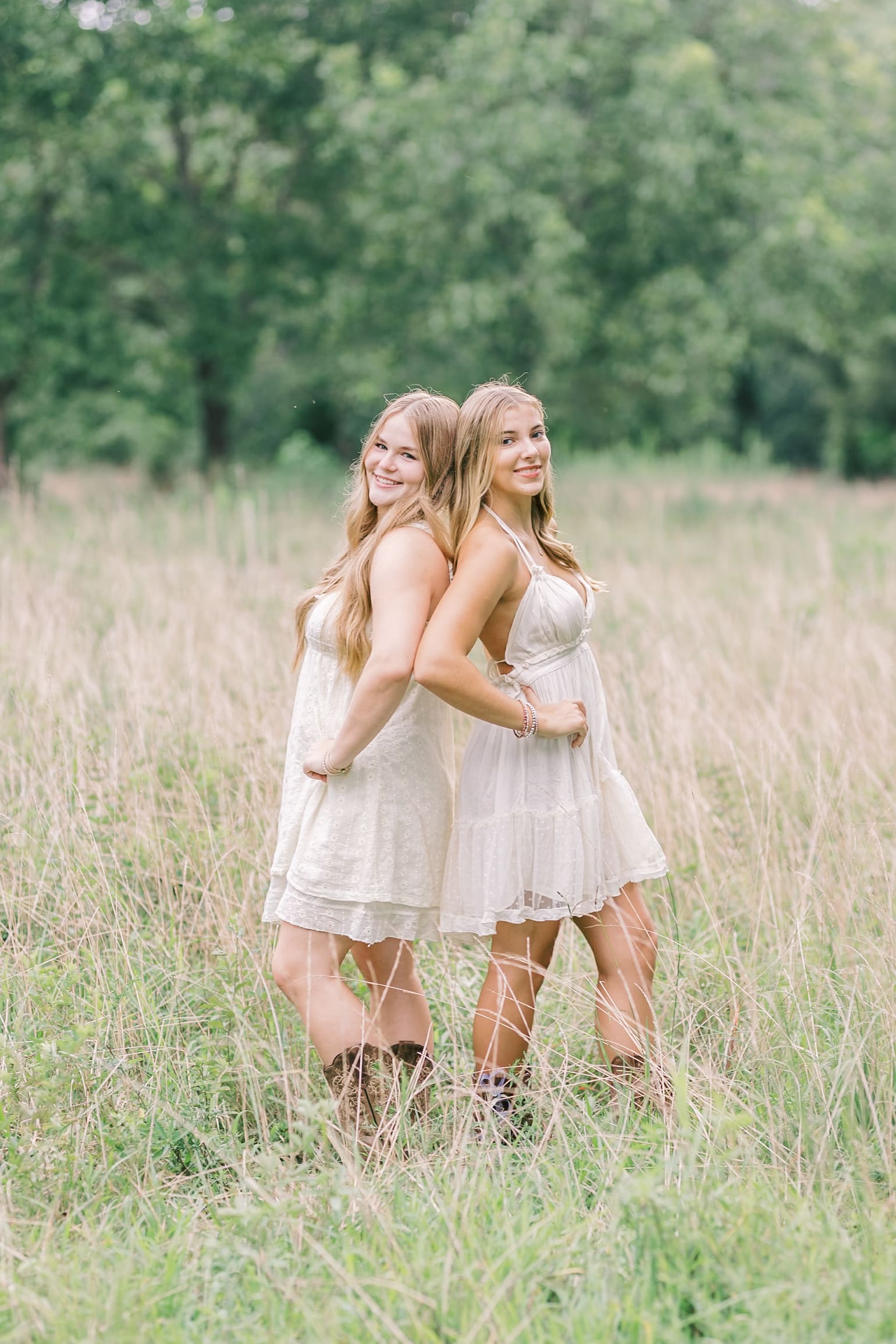 Best friend high school seniors lean back to back in white dresses in a pasture of tall grass