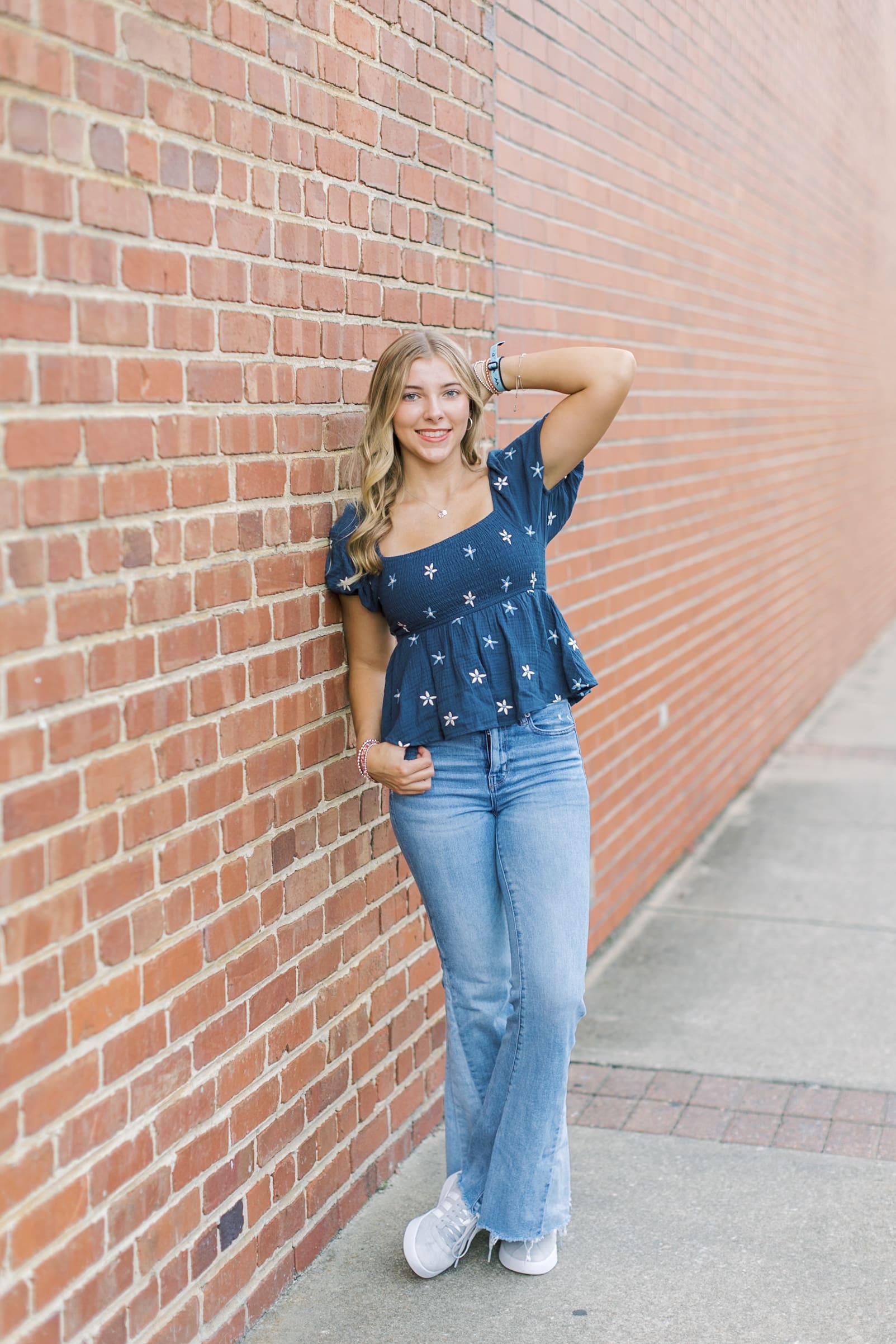 A blonde high school senior in jeans and a blue blouse from Raleigh boutiques leans against a brick wall with a hand in her hair