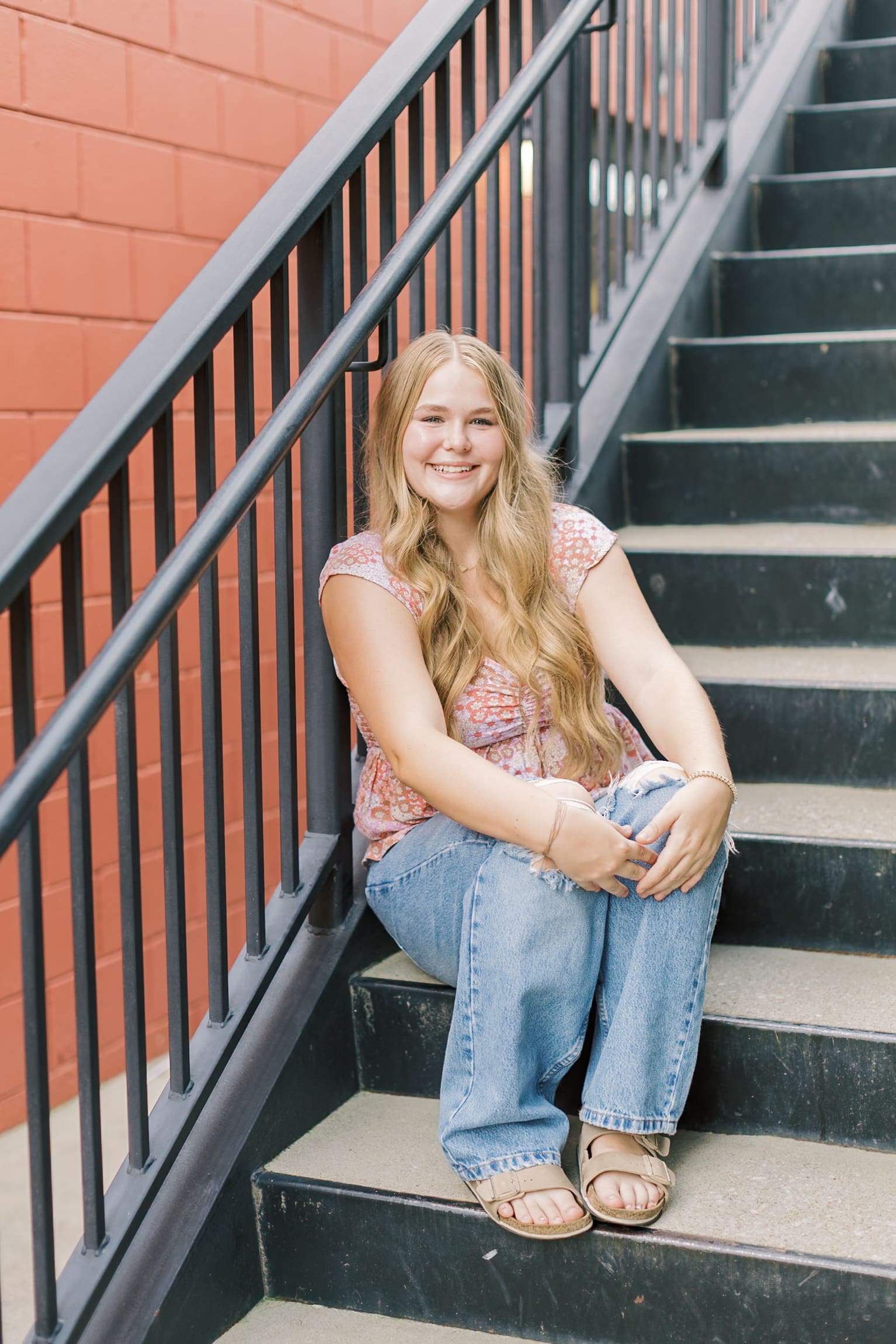 A happy high school senior smiles while sitting on outdoor stairs in a pink blouse and jeans after visiting Raleigh boutiques