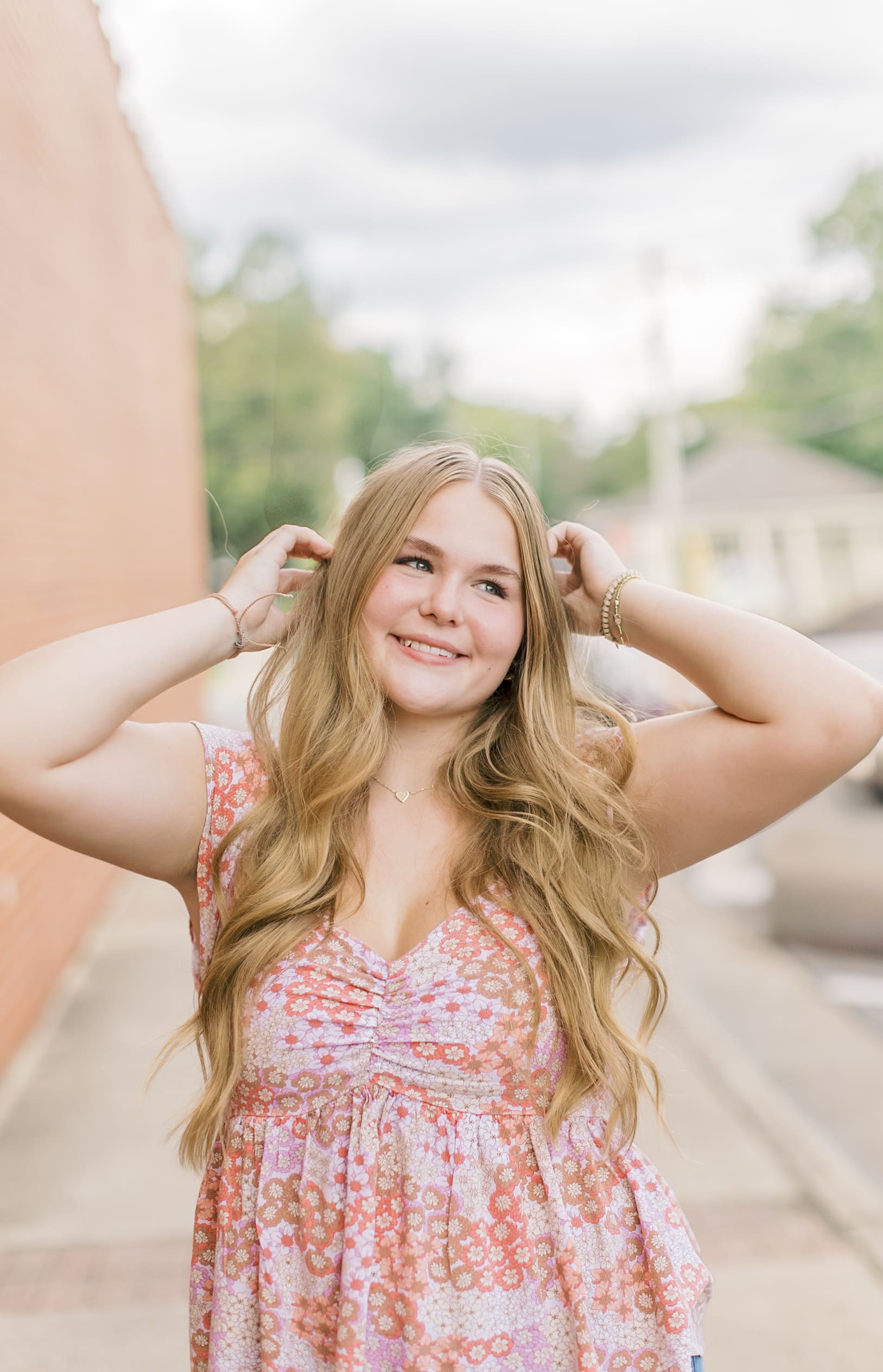 A happy high school senior in a pink floral print blouse pulls her hair back while walking a sidewalk