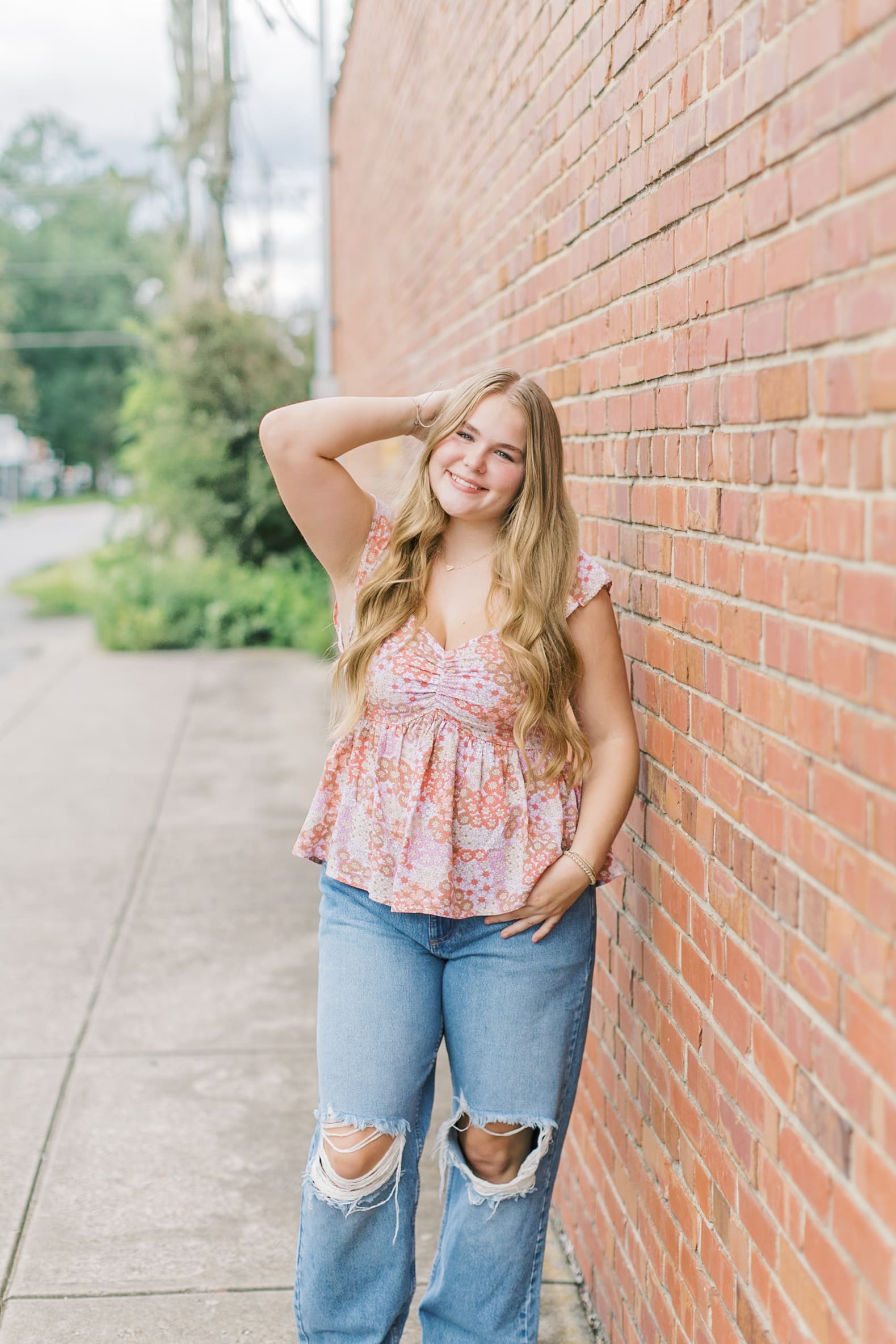 A happy teenager in a pink blouse and ripped jeans leans against a brick wall with a hand in her hair
