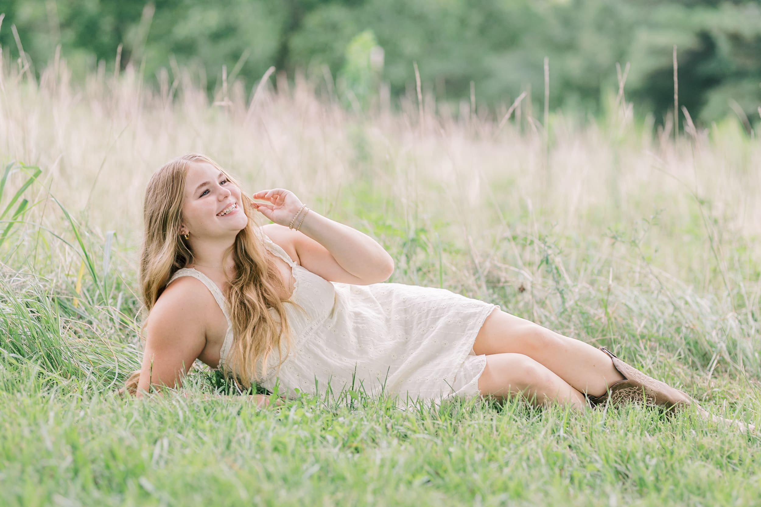 A laughing senior lays in some tall grass in a whtie dress and boots after visiting raleigh hair salons