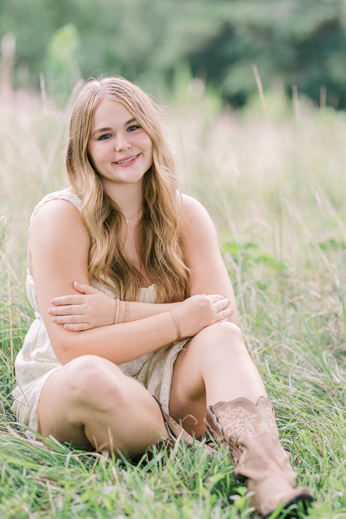 A happy teenager in a white dress and boots sits in tall grass with arms crossed and smiling after visiting raleigh hair salons
