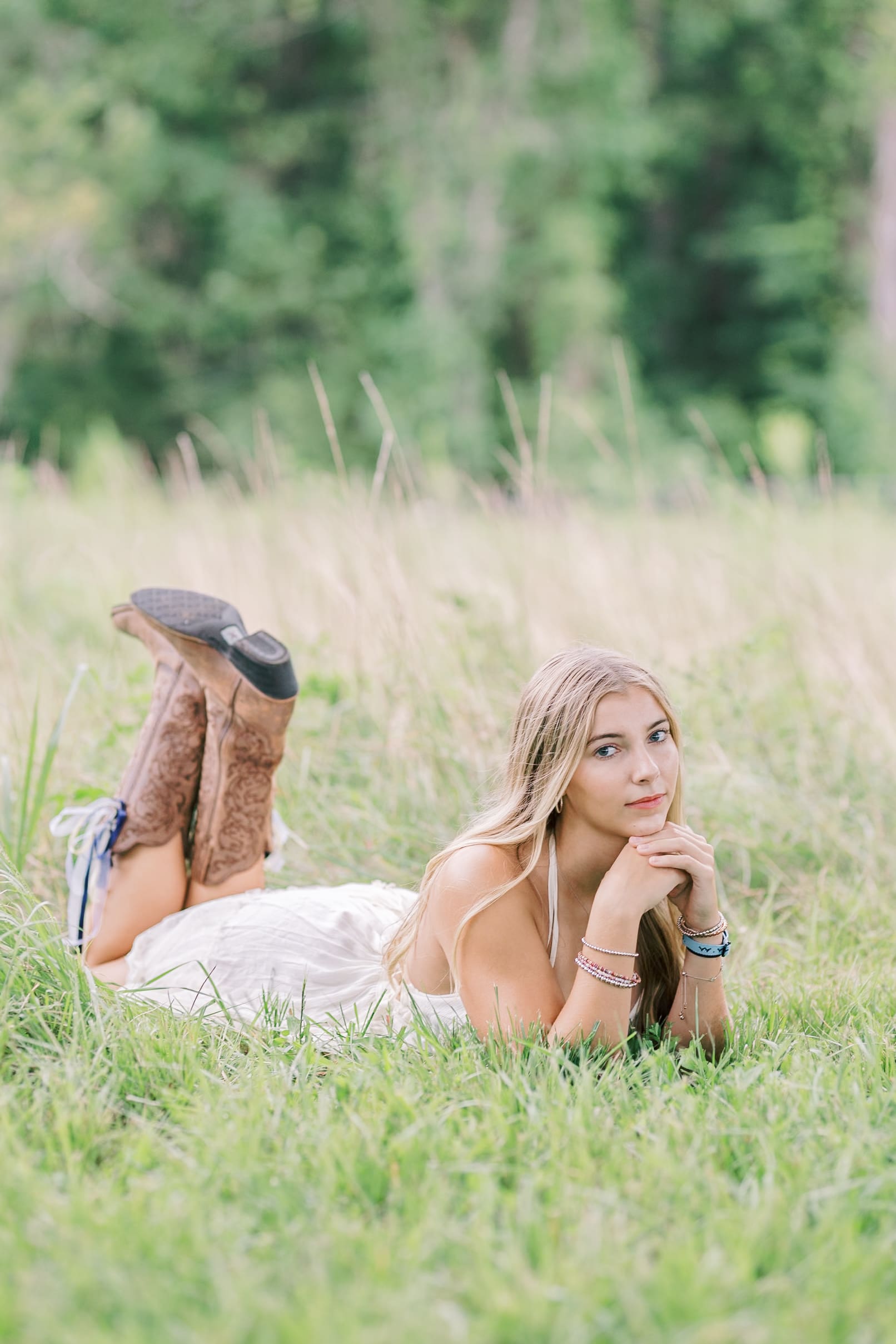 A blonde high school senior lays in the tall grass in a white dress leaning on her elbows with boots in the air behind her