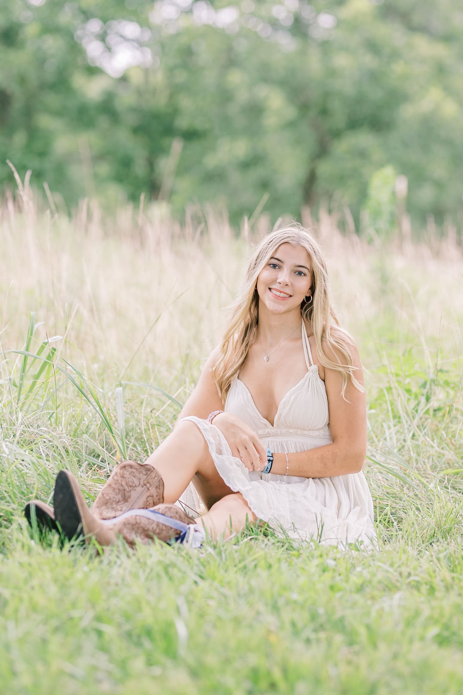 A high school senior in a white dress and boots sits in a field of tall grass after visiting Raleigh nail salons