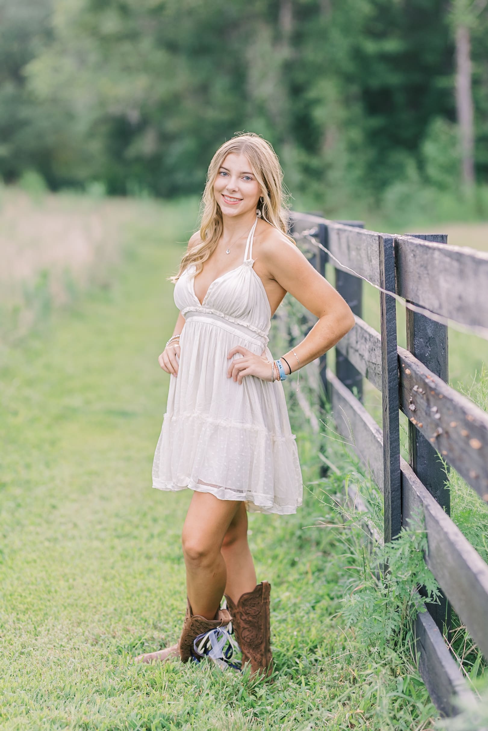 A high school grad in a white dress stands by a pasture fence in boots with hands on her hips