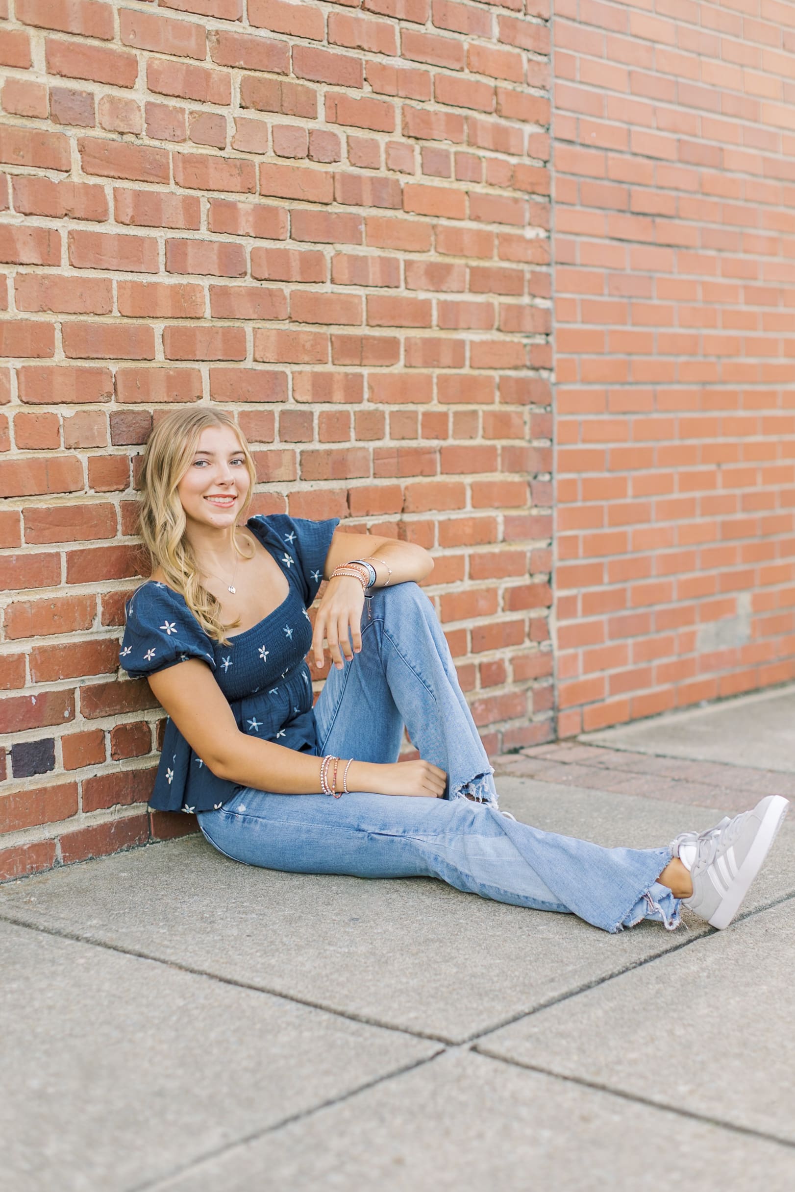 A blonde high school senior sits against a brick wall smiling in blue jeans and a blue blouse after visiting Raleigh nail salons