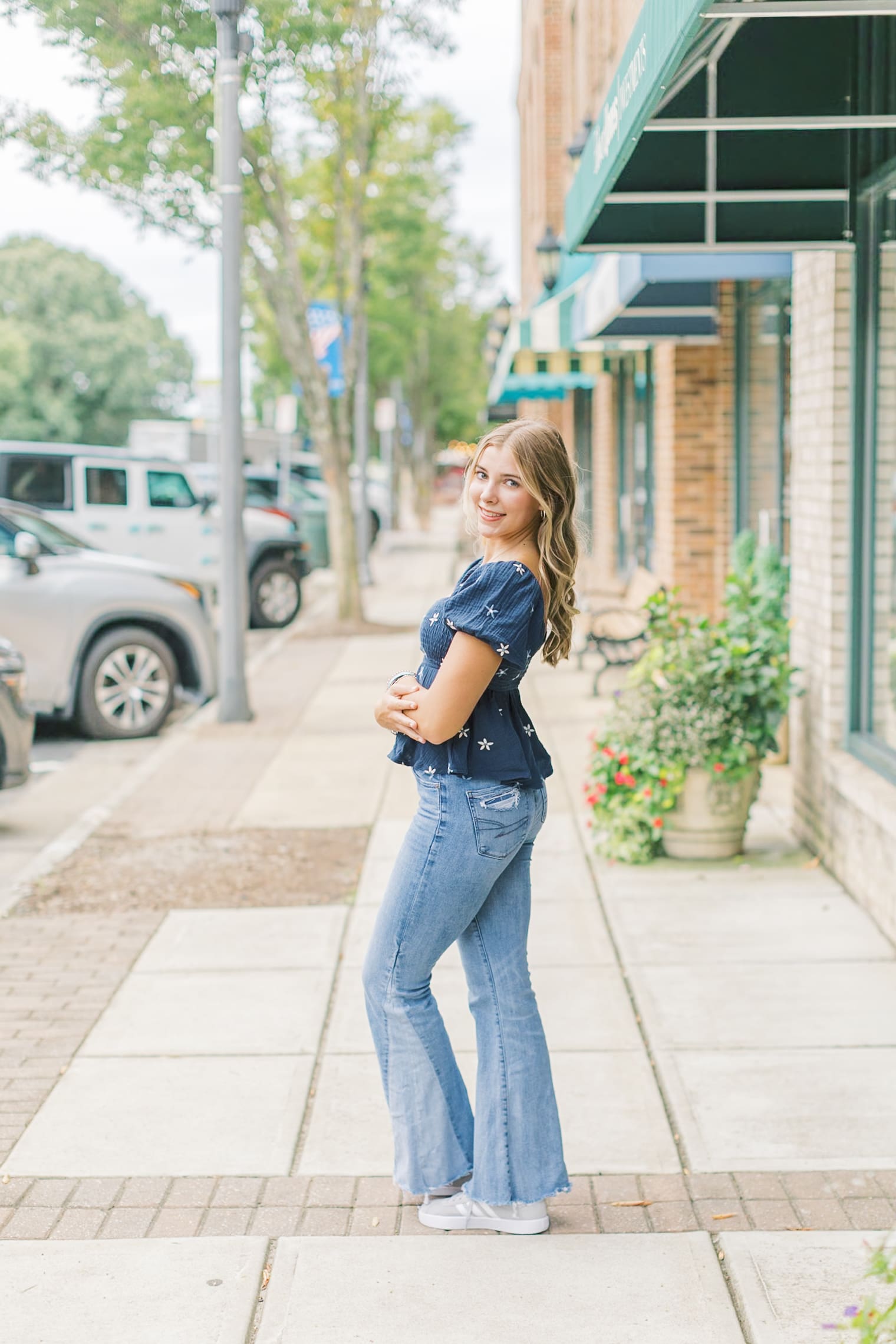 A high school senior smiles over her shoulder while walking down a sidewalk in a blue blouse and jeans after visiting Raleigh nail salons