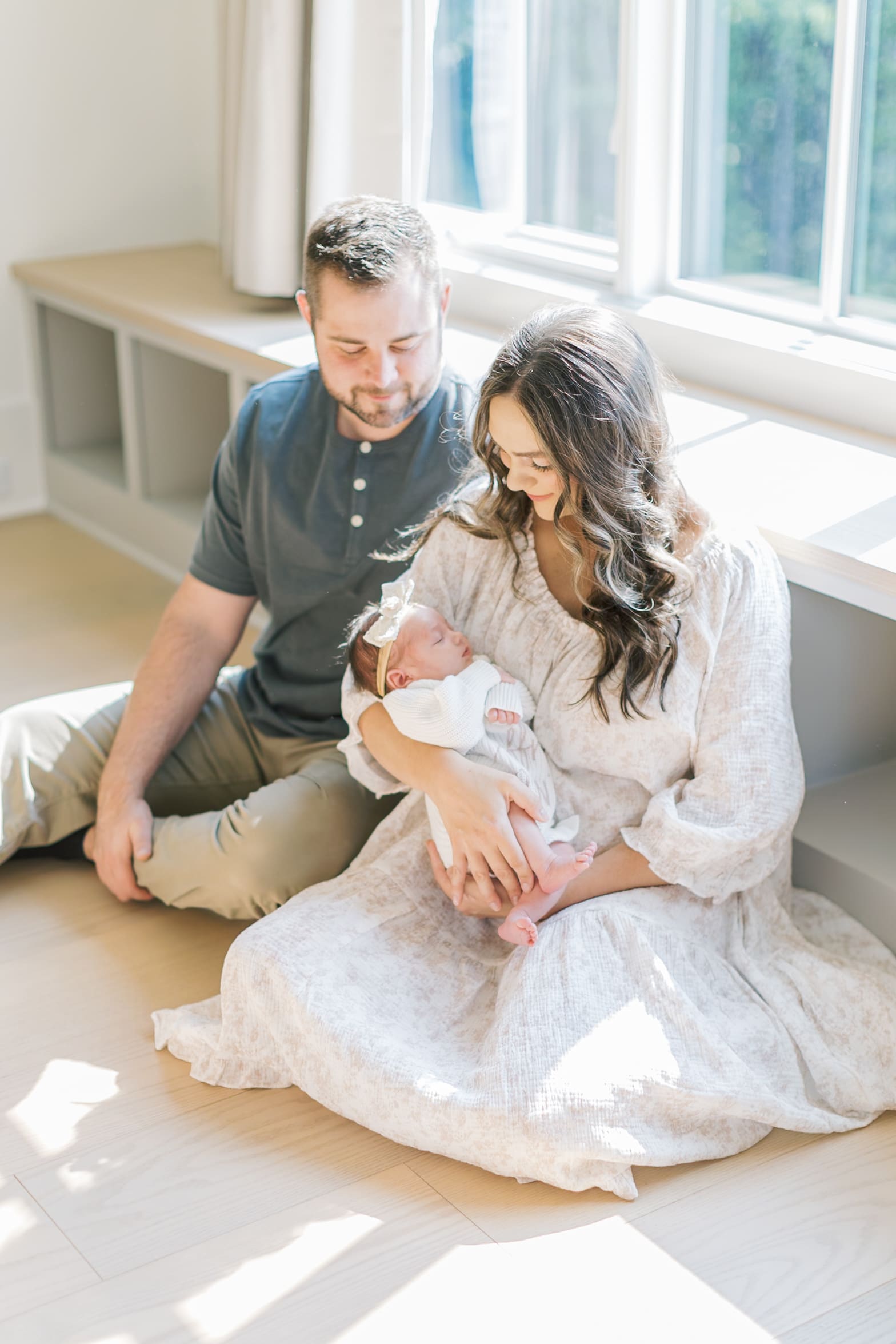 Mom and dad sit on the floor under a window cradling their sleeping newborn daughter