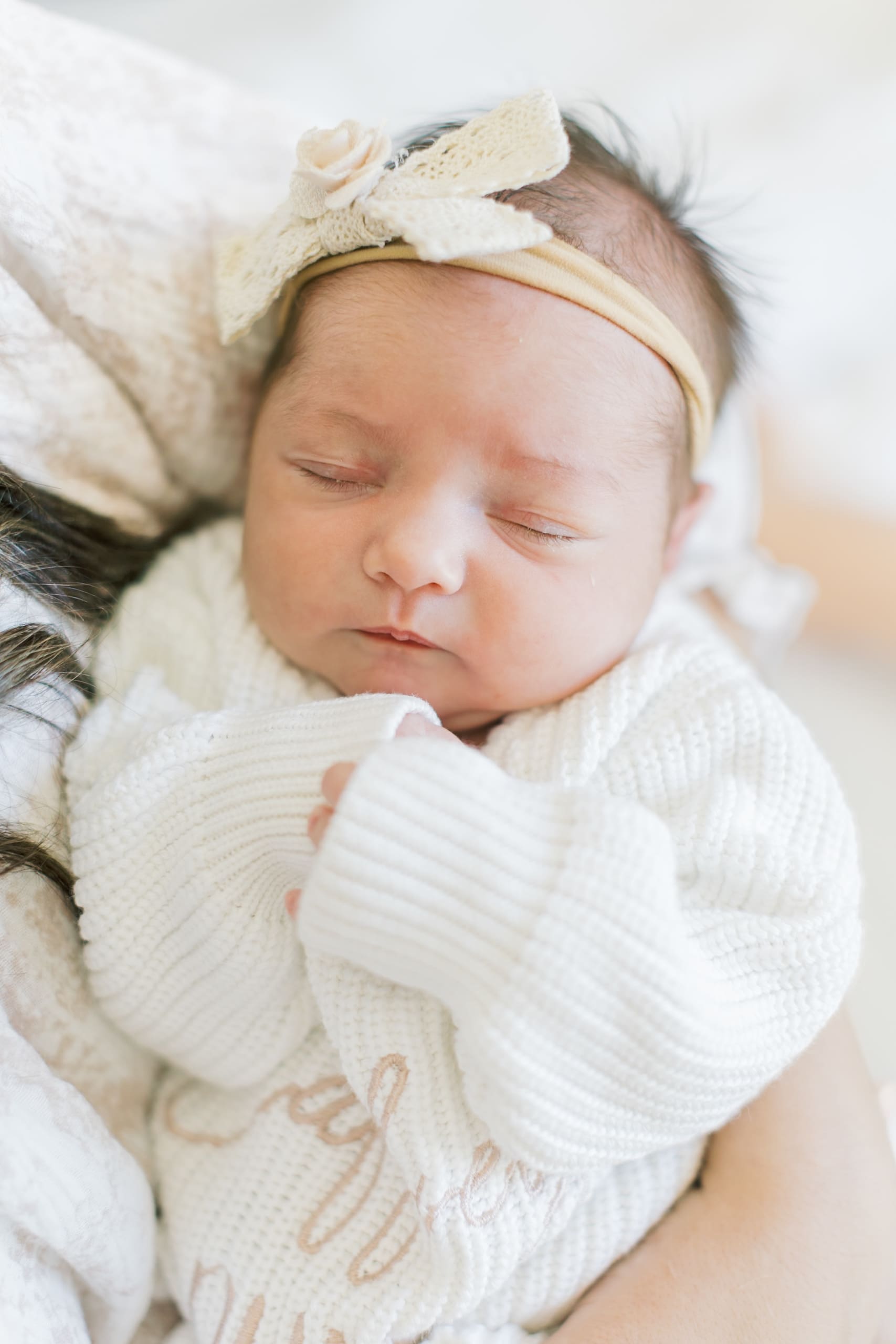 A newborn baby in a white sweater and bow sleeps in mom's arms