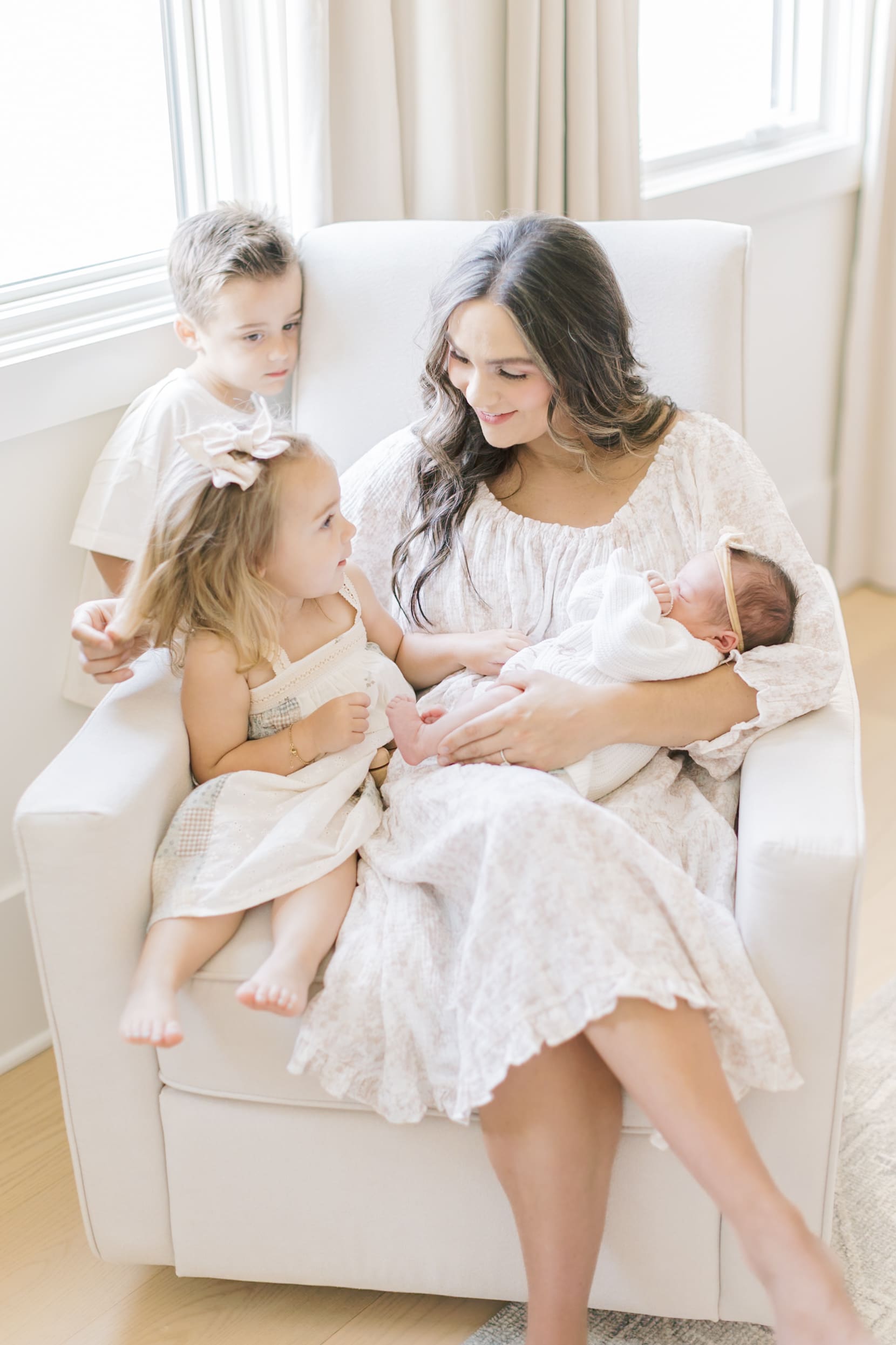 A happy mom in a white dress sits in a nursing chair with her newborn in her lap and toddler son and daughter at her side after meeting Raleigh nannies