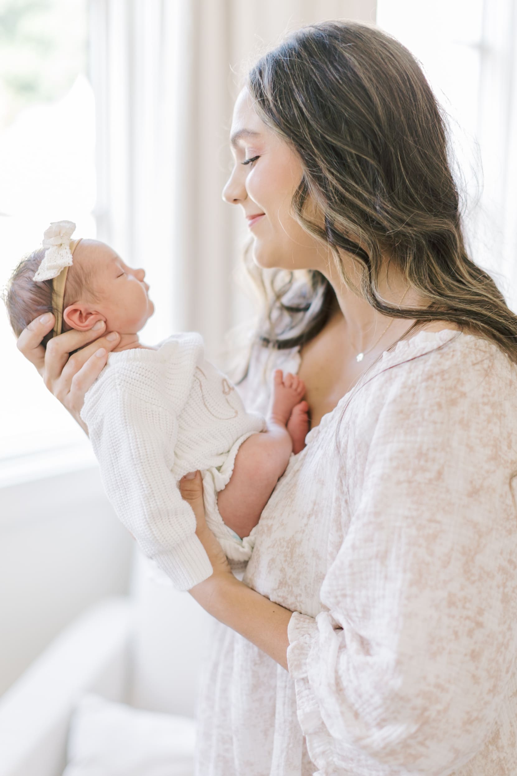 A happy new mom in a white gown smiles down at her sleeping newborn in her hands after meeting Raleigh nannies