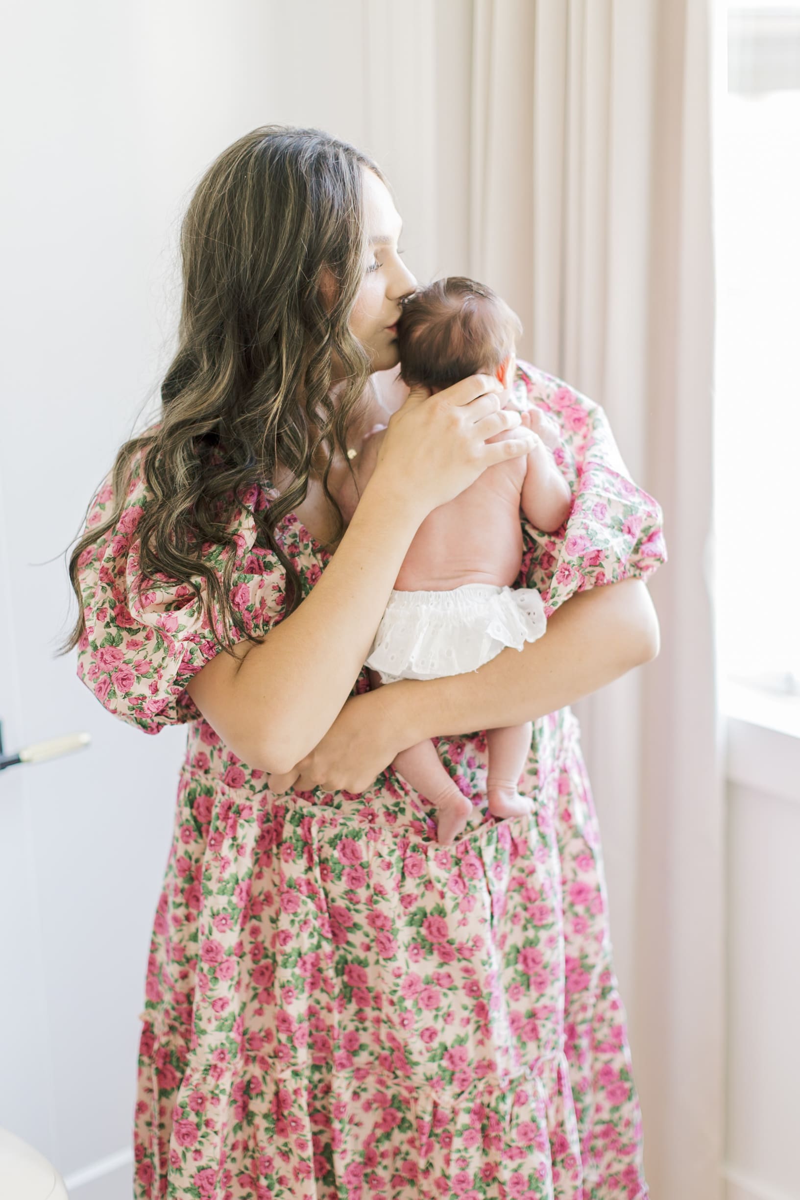 A new mom in a pink flower print dress kisses her newborn while holding it against her chest and standing in a window after meeting Raleigh nannies