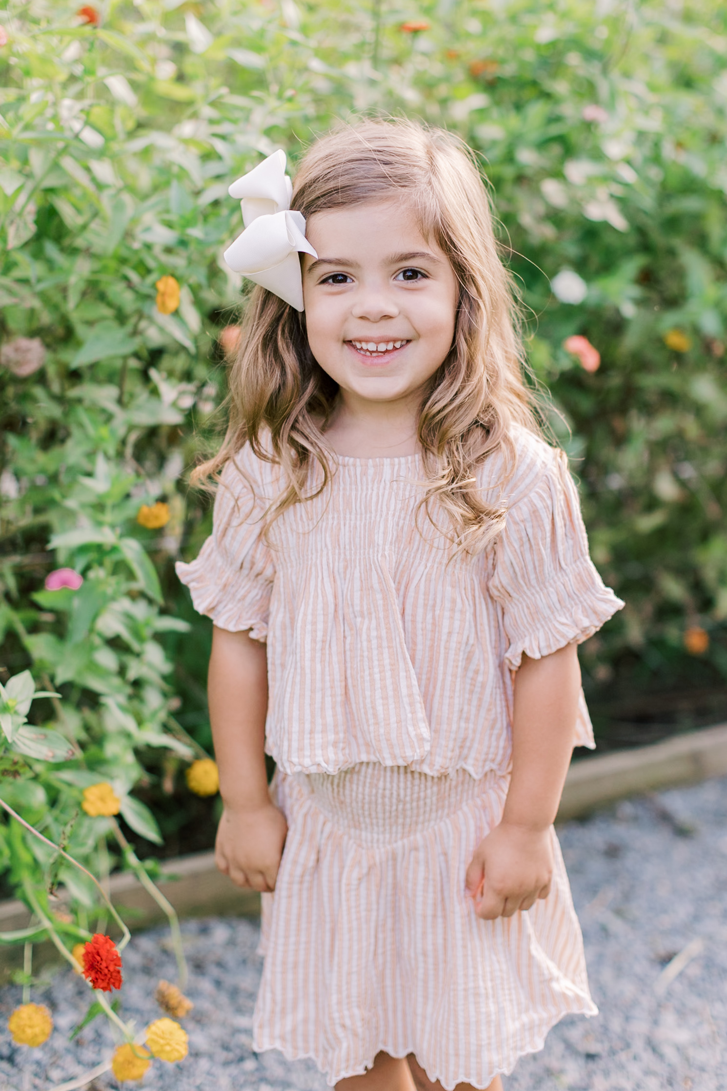 A happy toddler girl in a pink stripe dress standing in a garden with flowers