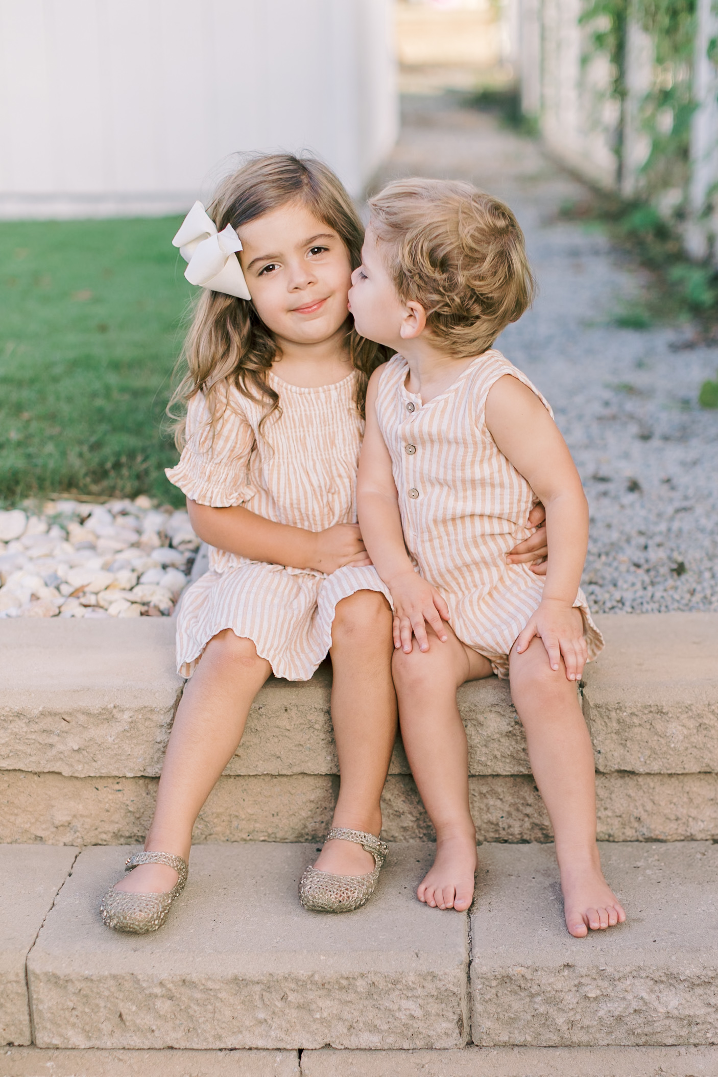 A young toddler boy kisses the cheek of his slightly older sister as they sit on stone garden steps in matching outfits after visiting daycares in raleigh