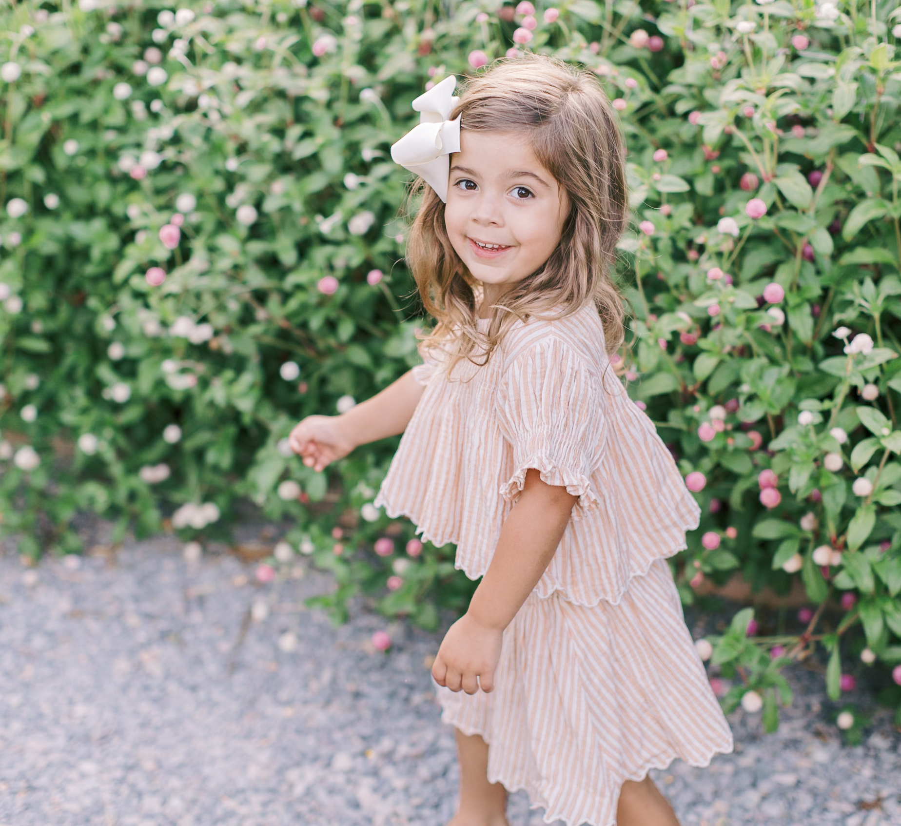 A toddler girl in a pink dress leads the way through a garden with a big smile after visiting daycares in raleigh