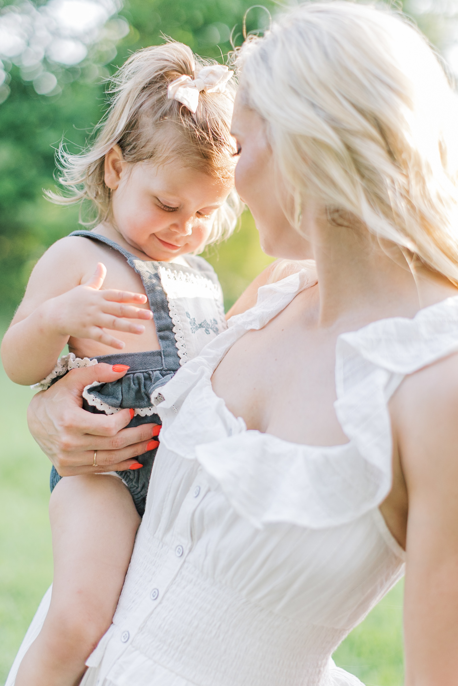 A toddler girl sits on mom's hip in a denim overalls dress in a park at sunset