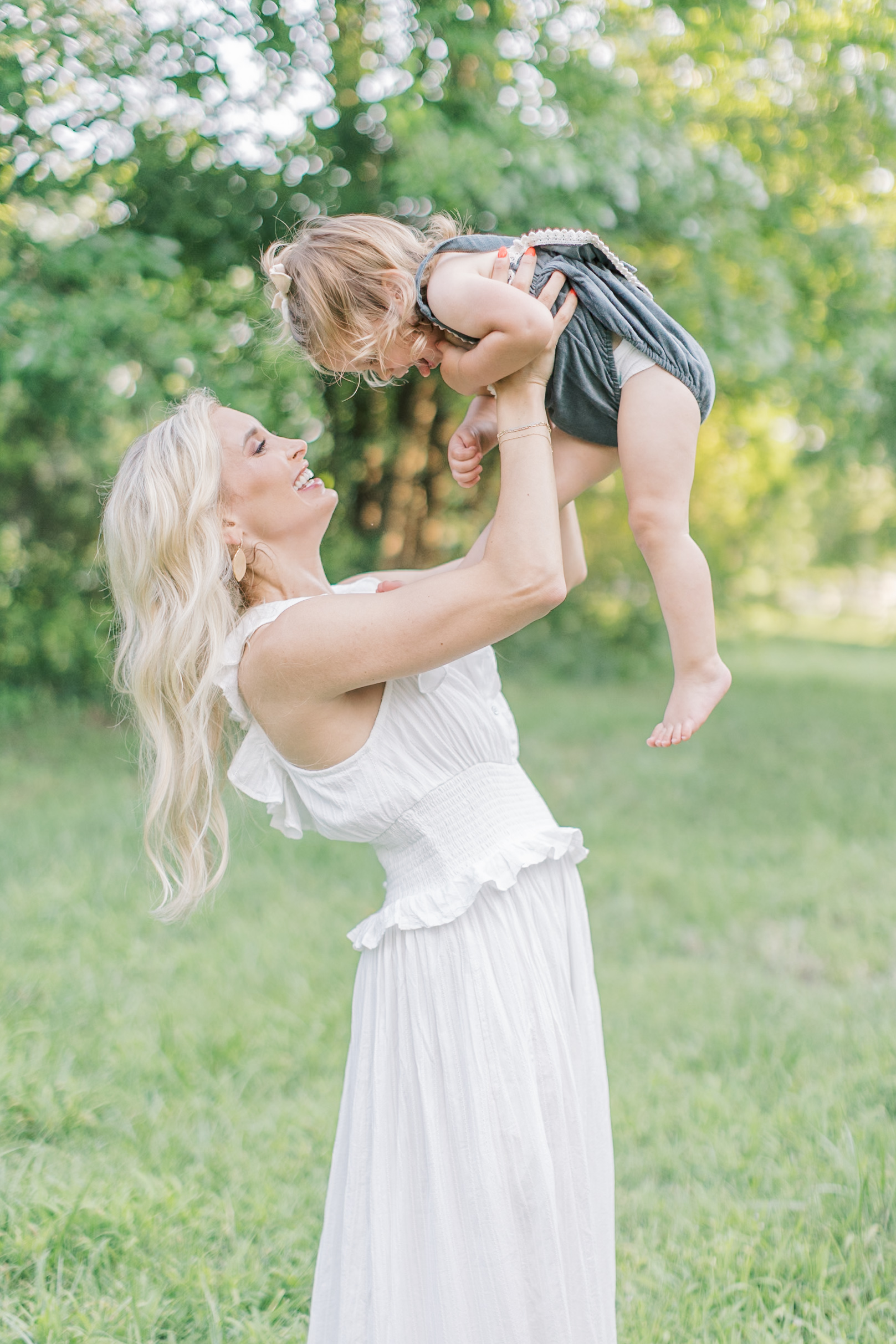 A happy mom in a white dress lifts her toddler daughter over her head in a park at sunset after some mommy and me classes in raleigh