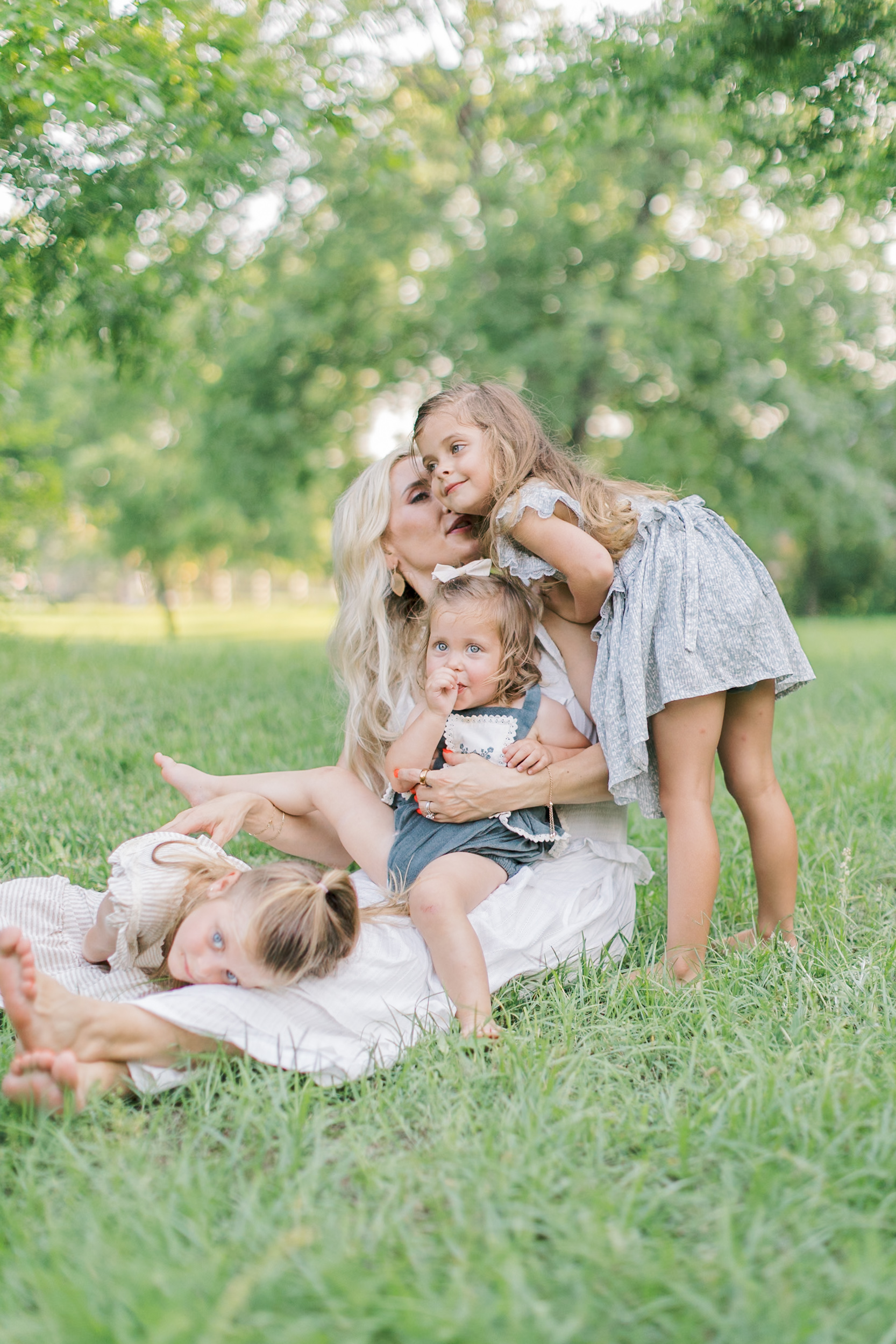 Three toddler sisters play in mom's lap in a lawn with green grass in dresses after some mommy and me classes in raleigh