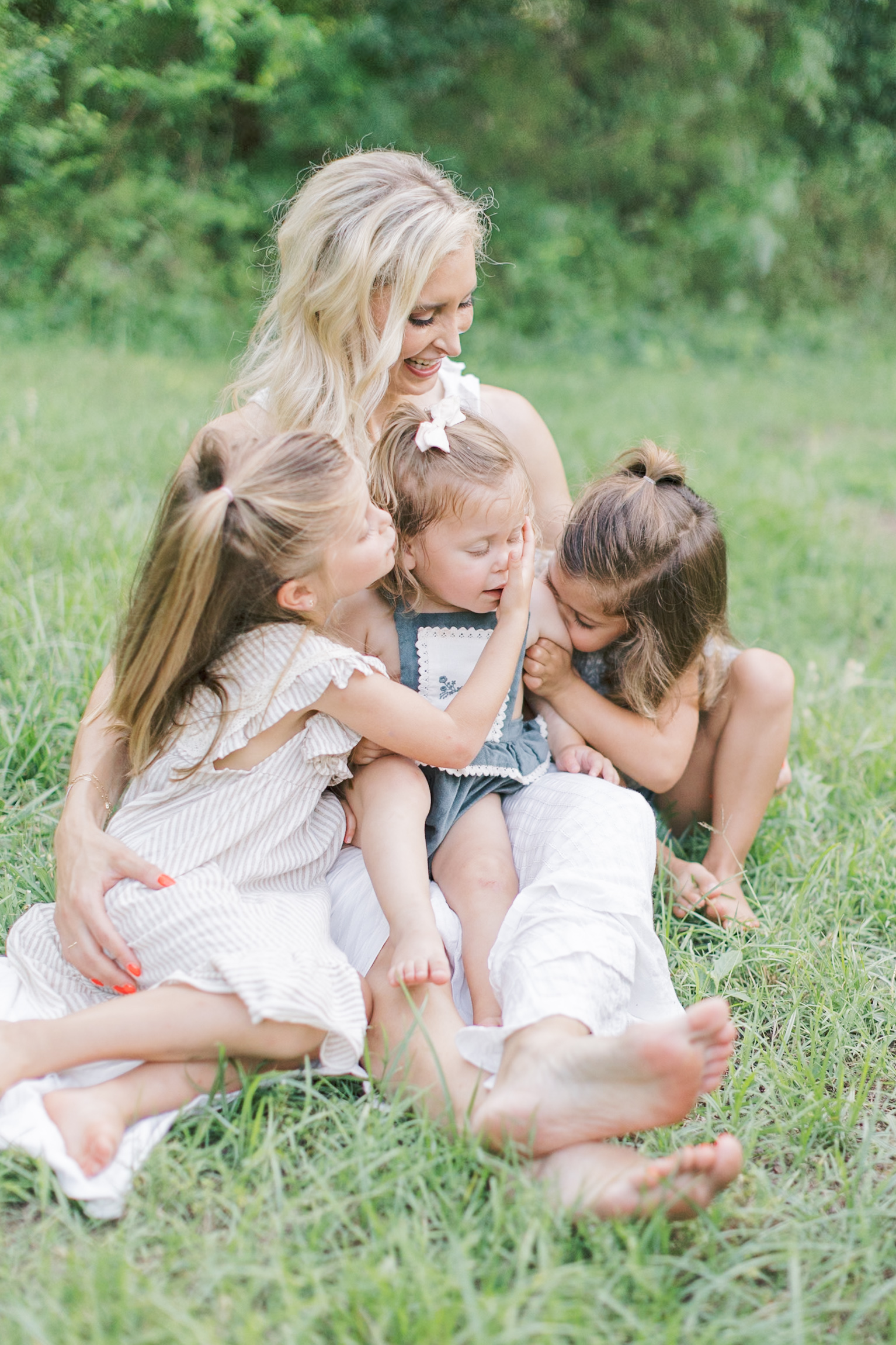 A laughing mother sits in the grass with her three young daughters playing in her lap after some mommy and me classes in raleigh