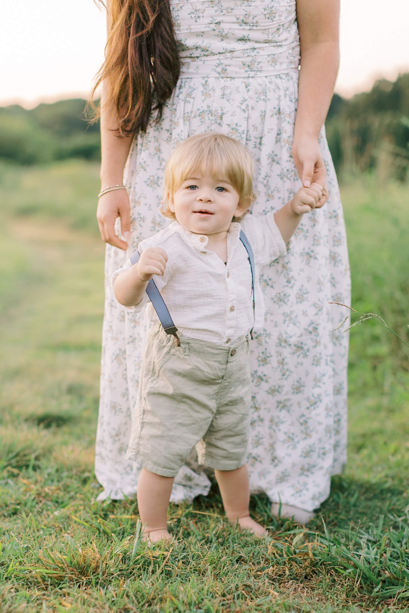 A young boy walks in front of mom holding her hand in suspenders and white shirt