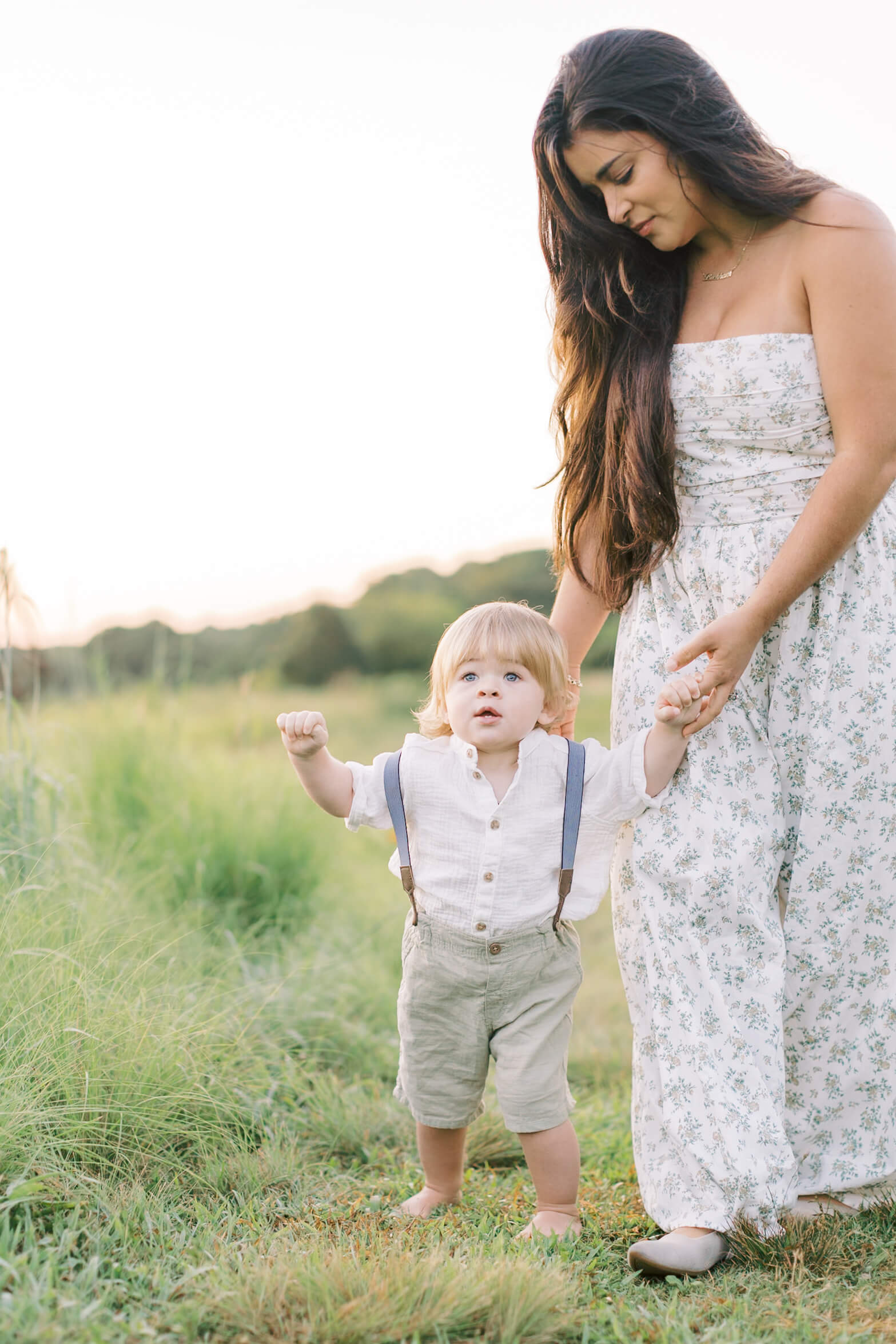 A young toddler in suspenders walks with mom's help in a field of tall grass at sunset