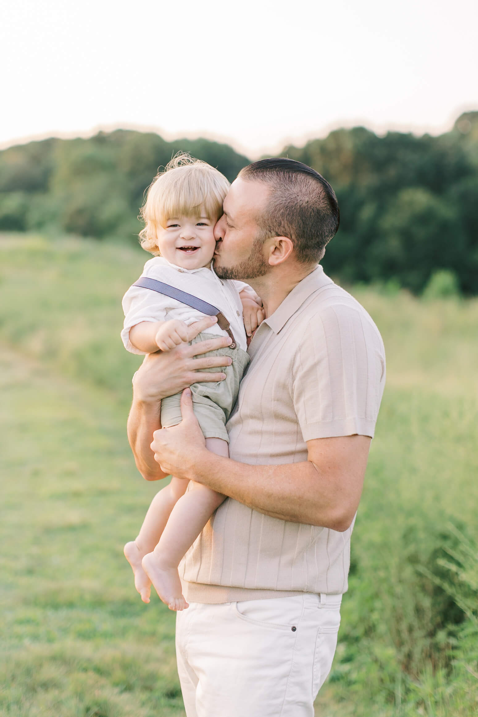 A young boy in suspenders and white shirt smiles while being kissed by dad in a field at sunset after meeting pediatricians in raleigh