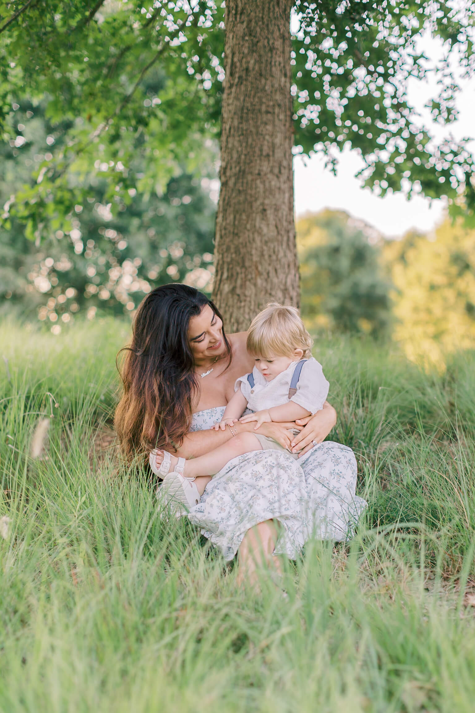 A happy mom in a flower dress sits under a tree in tall grass playing with her infant in her lap after meeting pediatricians in raleigh